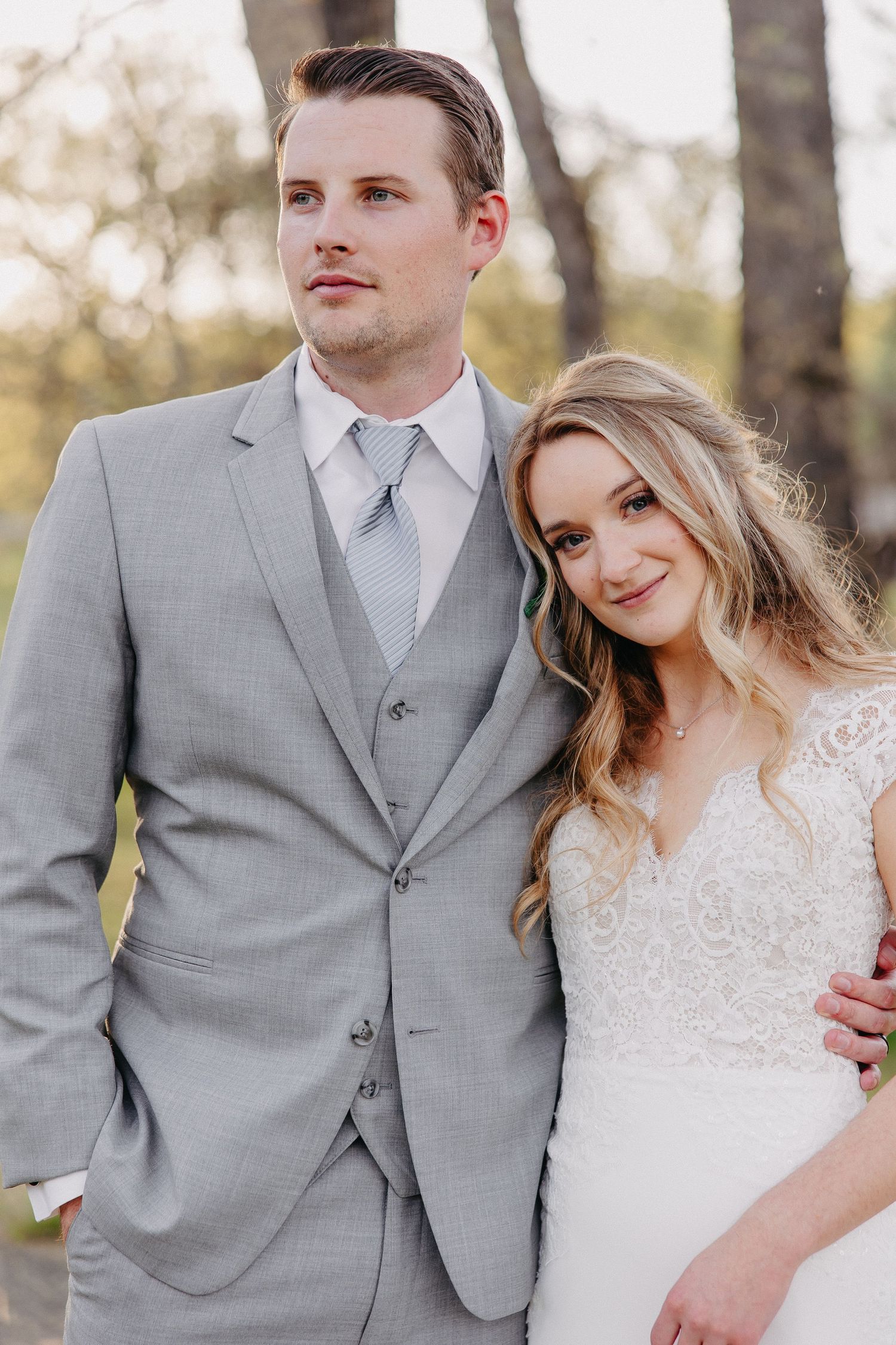 Portrait of newlyweds in gray suit and lace dress smiling together outdoors.