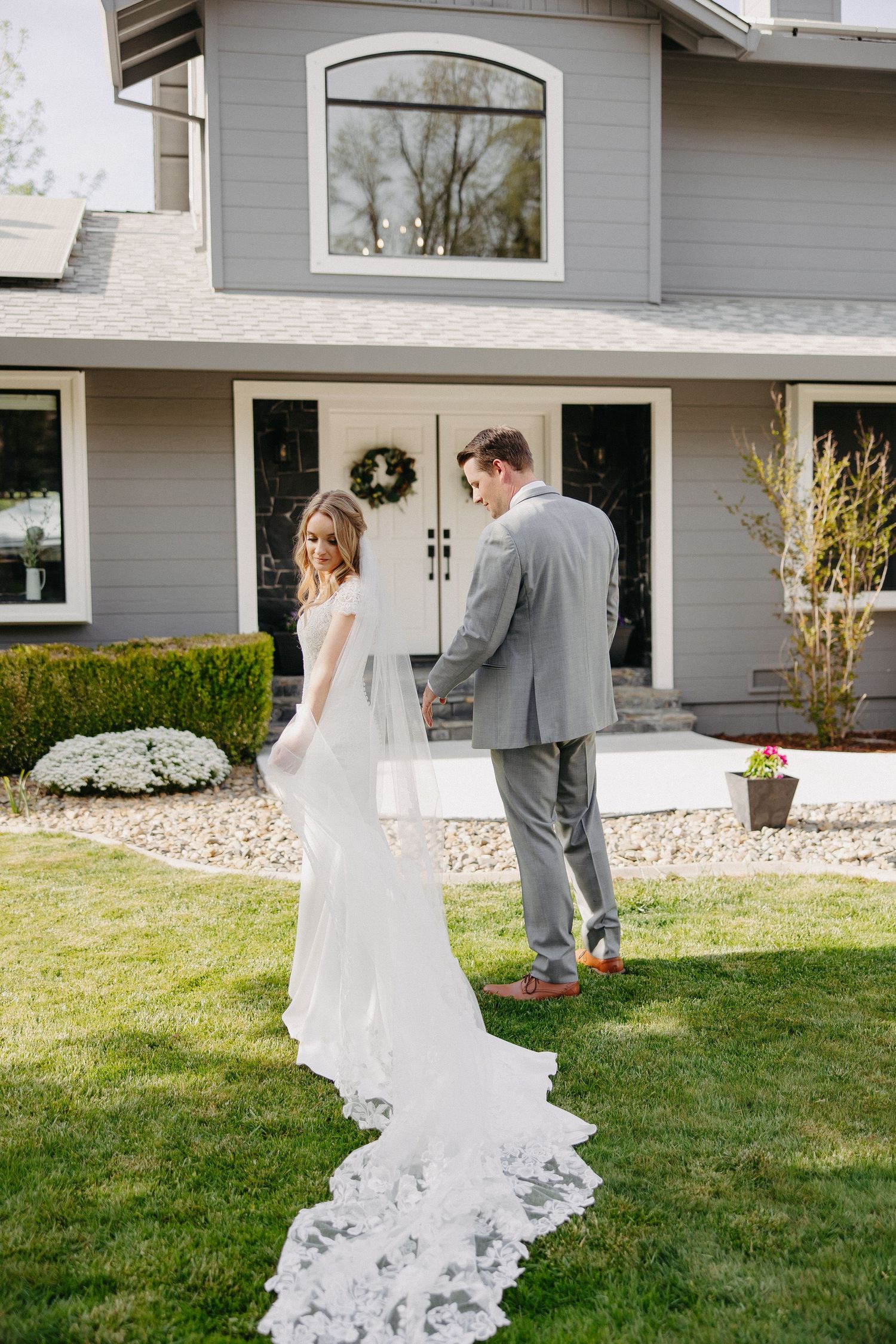 A wedding couple stands in front of a gray house with a beautiful lace train on the bride's dress.