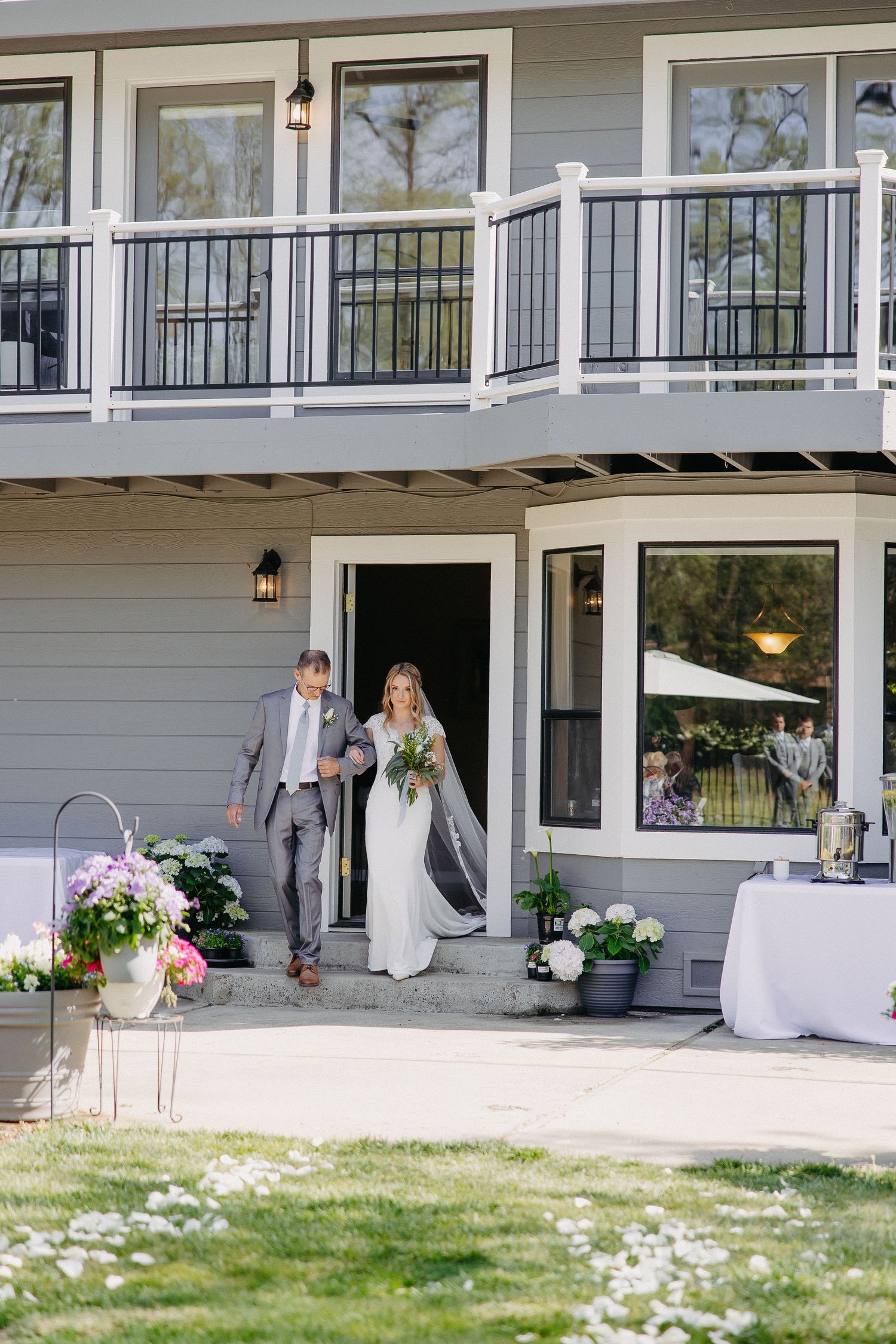 A couple emerges from a grey house with balcony during an outdoor wedding celebration.