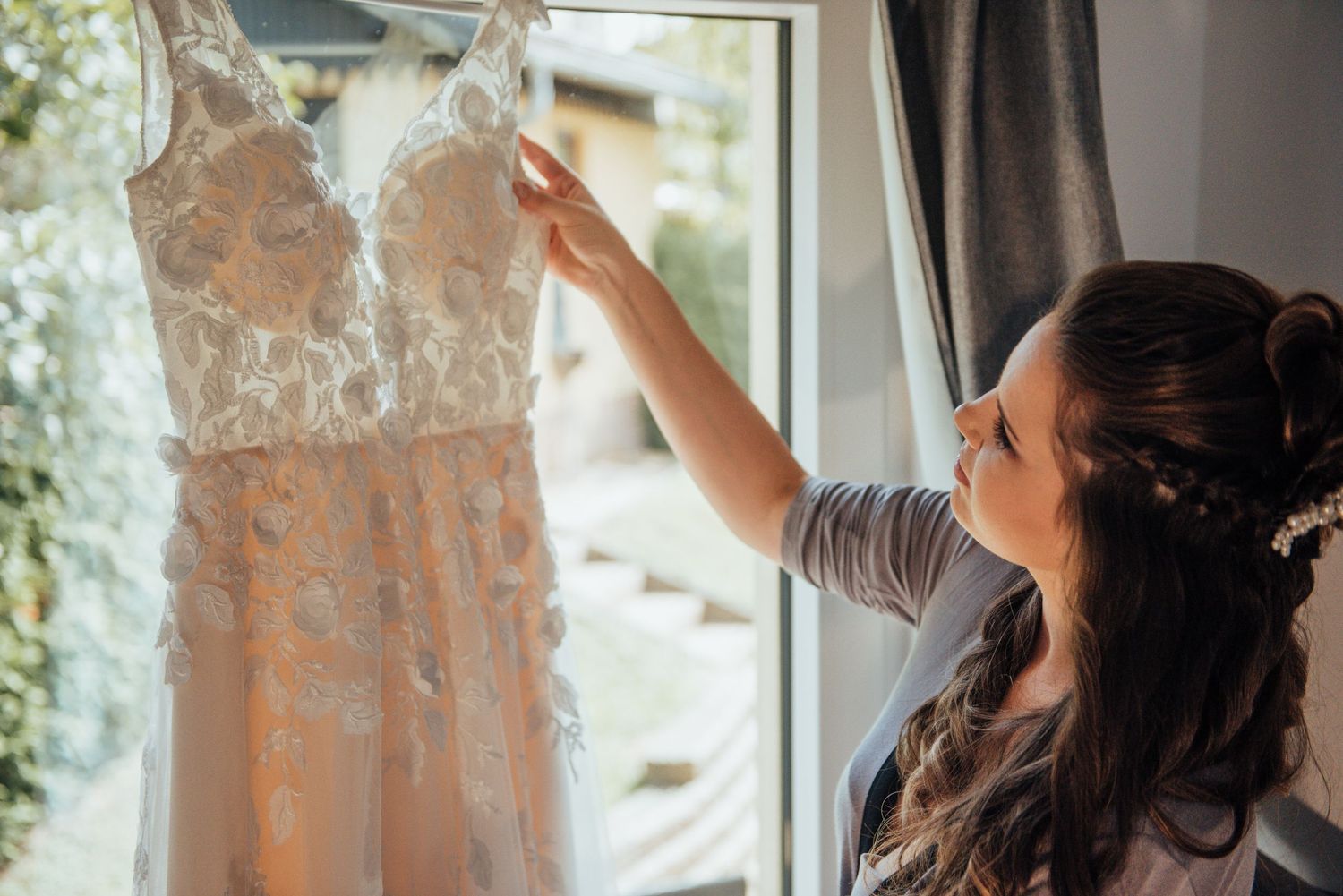 A delicate lace wedding dress hangs in window light as someone reaches up to touch it.