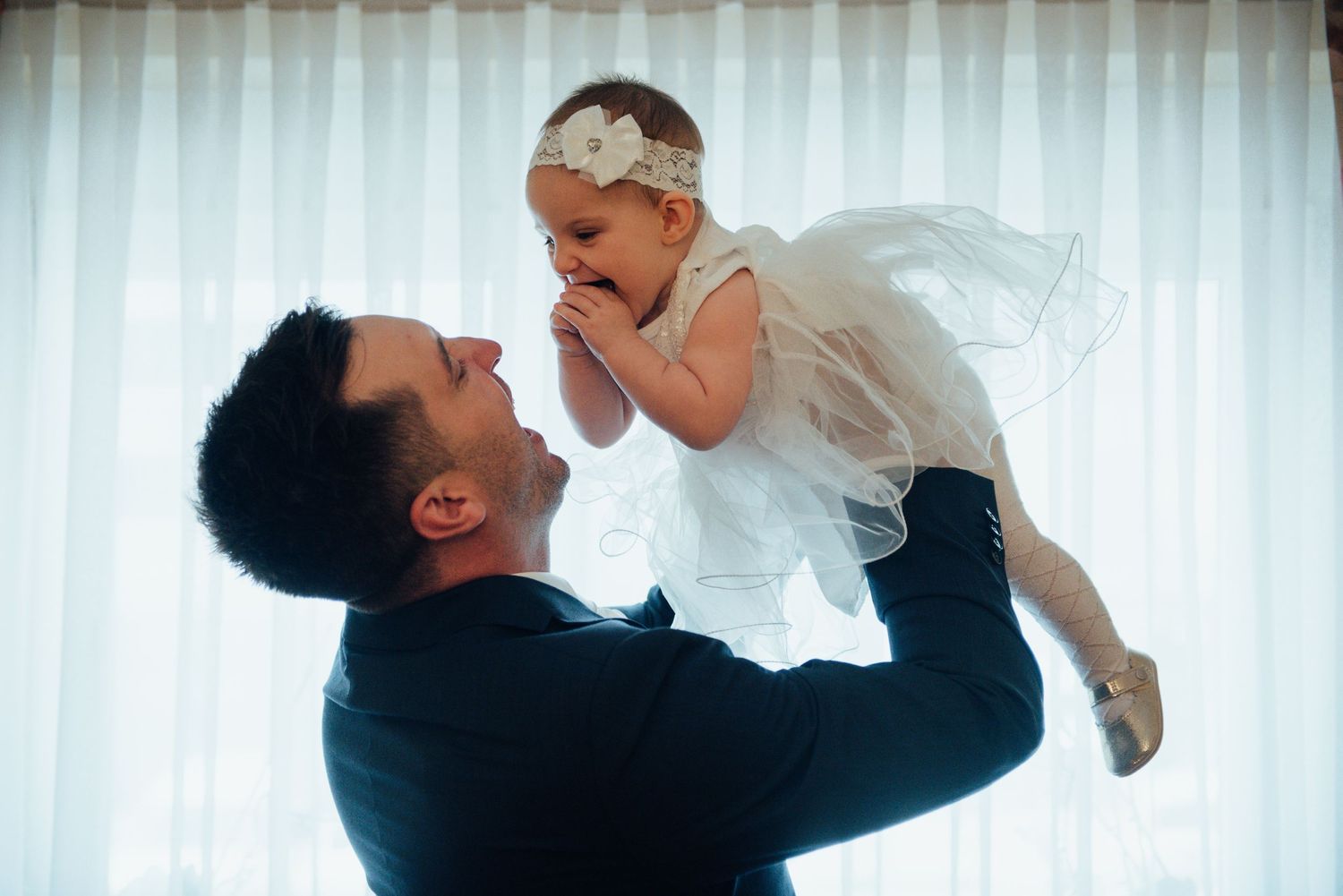 A joyful moment between parent and child in formal wear against a bright window.