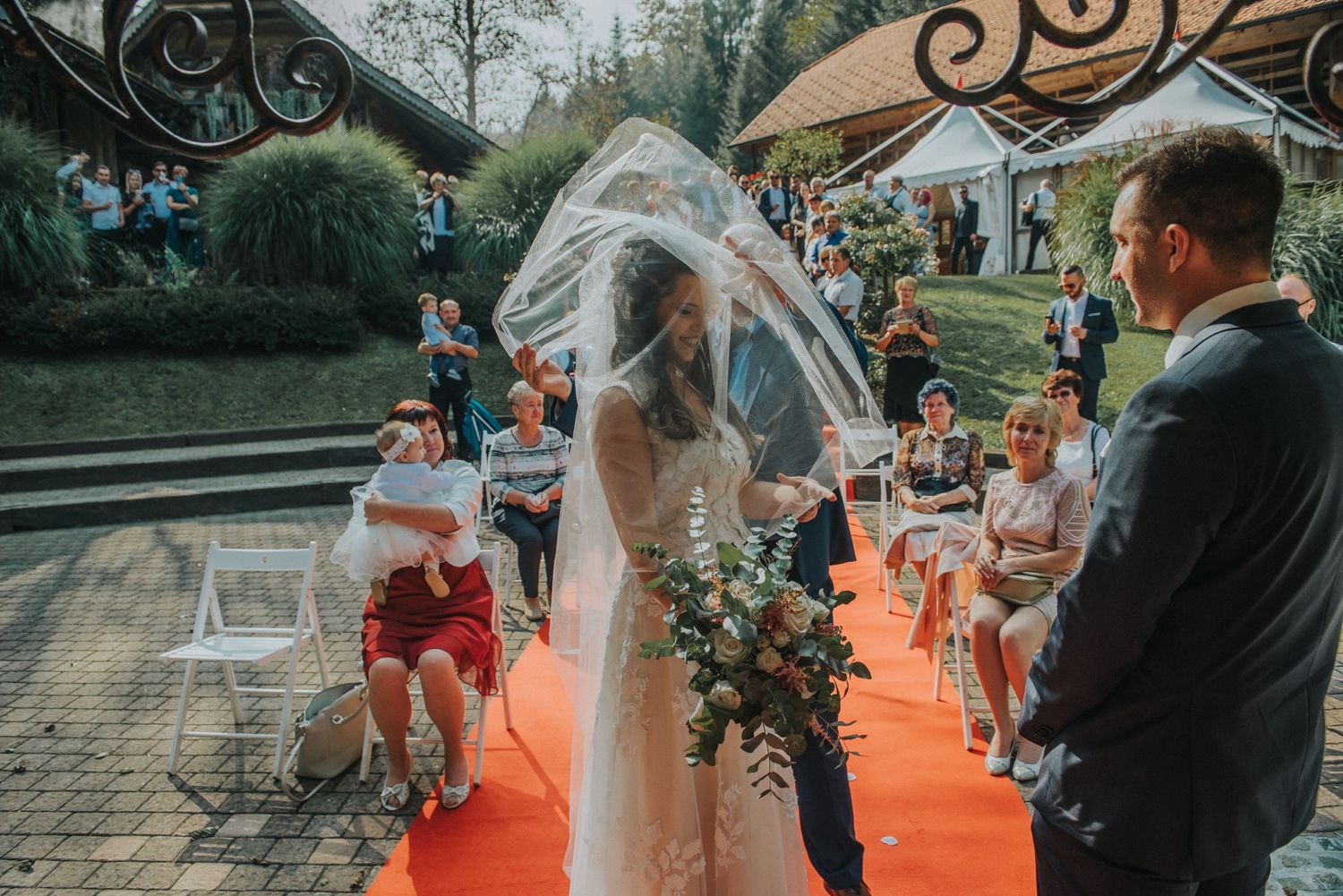 An outdoor wedding ceremony with a red carpet aisle and decorative arch.