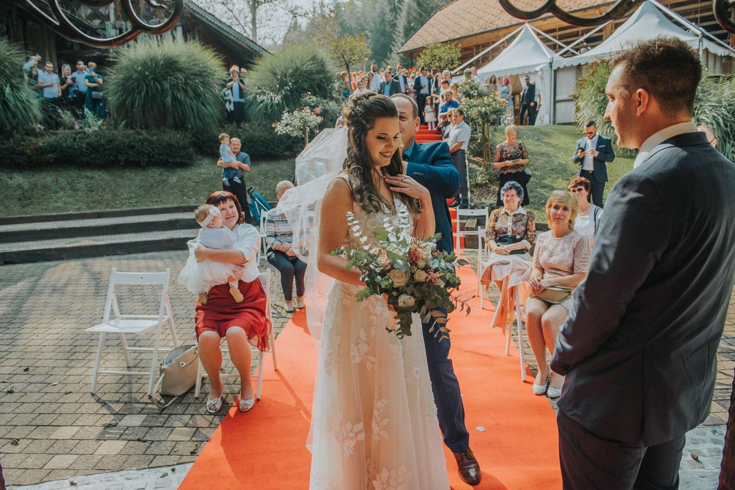 A wedding ceremony taking place on a red carpet aisle with guests seated outdoors.