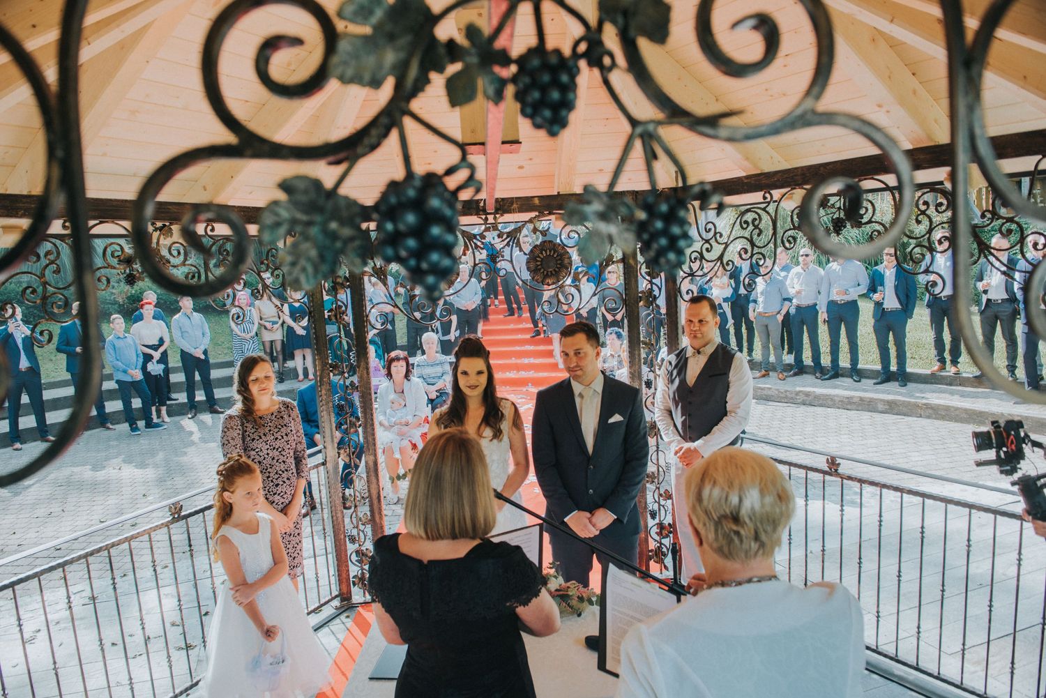 A wedding ceremony takes place under an ornate gazebo decorated with floral arrangements.