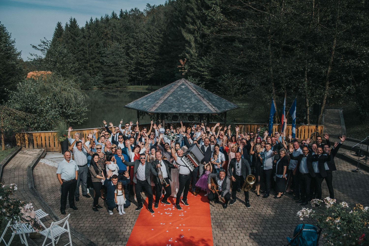 A large group of wedding guests poses for a photo in front of a forest gazebo with red carpet.