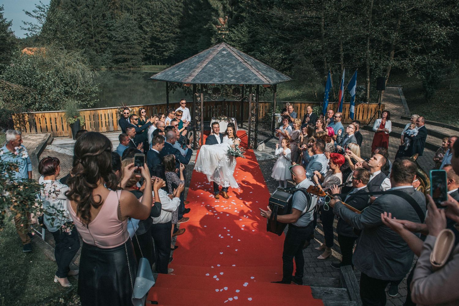 Wedding guests celebrate and throw petals as newlyweds walk down a red carpet outdoor aisle.