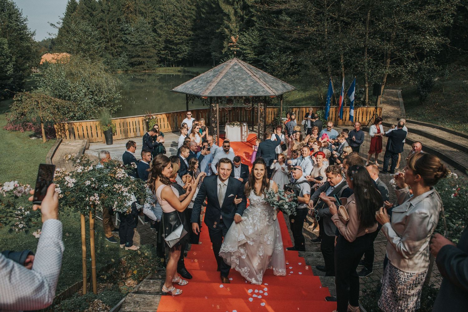 Wedding guests line a red carpet path throwing petals during an outdoor celebration.
