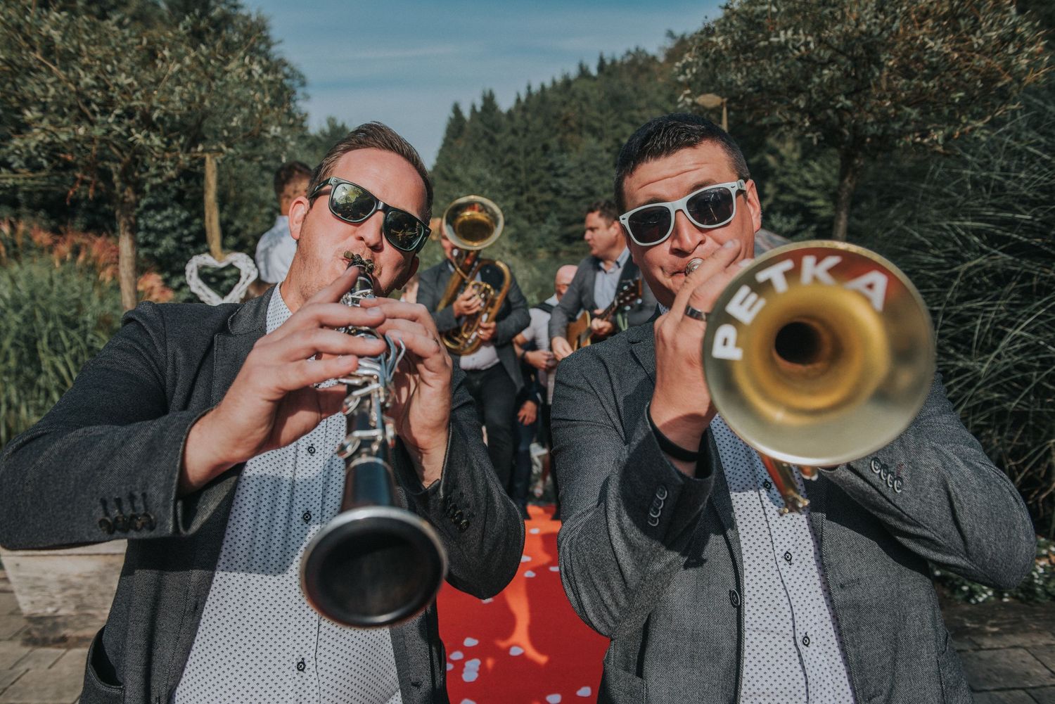 Two musicians playing brass instruments perform outdoors at a wedding celebration.