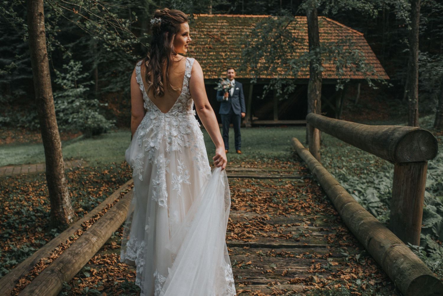 Elegant lace wedding dress photographed from behind on woodland path with rustic cabin in background.