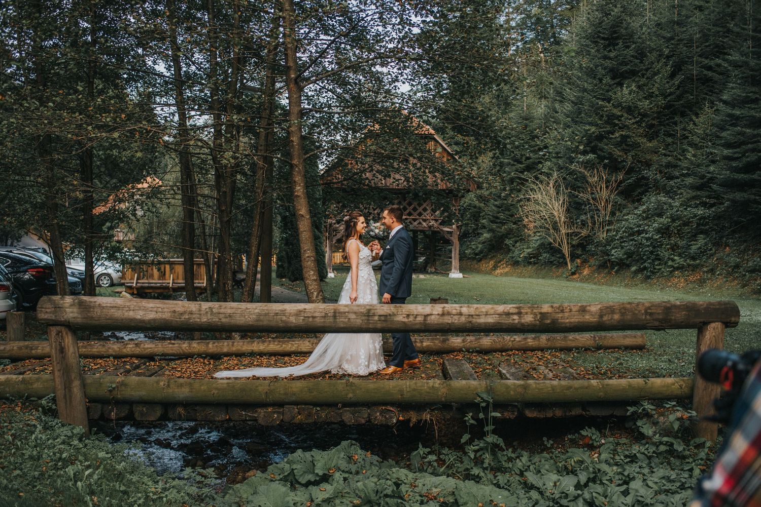 Wedding couple poses on wooden fence with rustic cabin and evergreen forest backdrop.