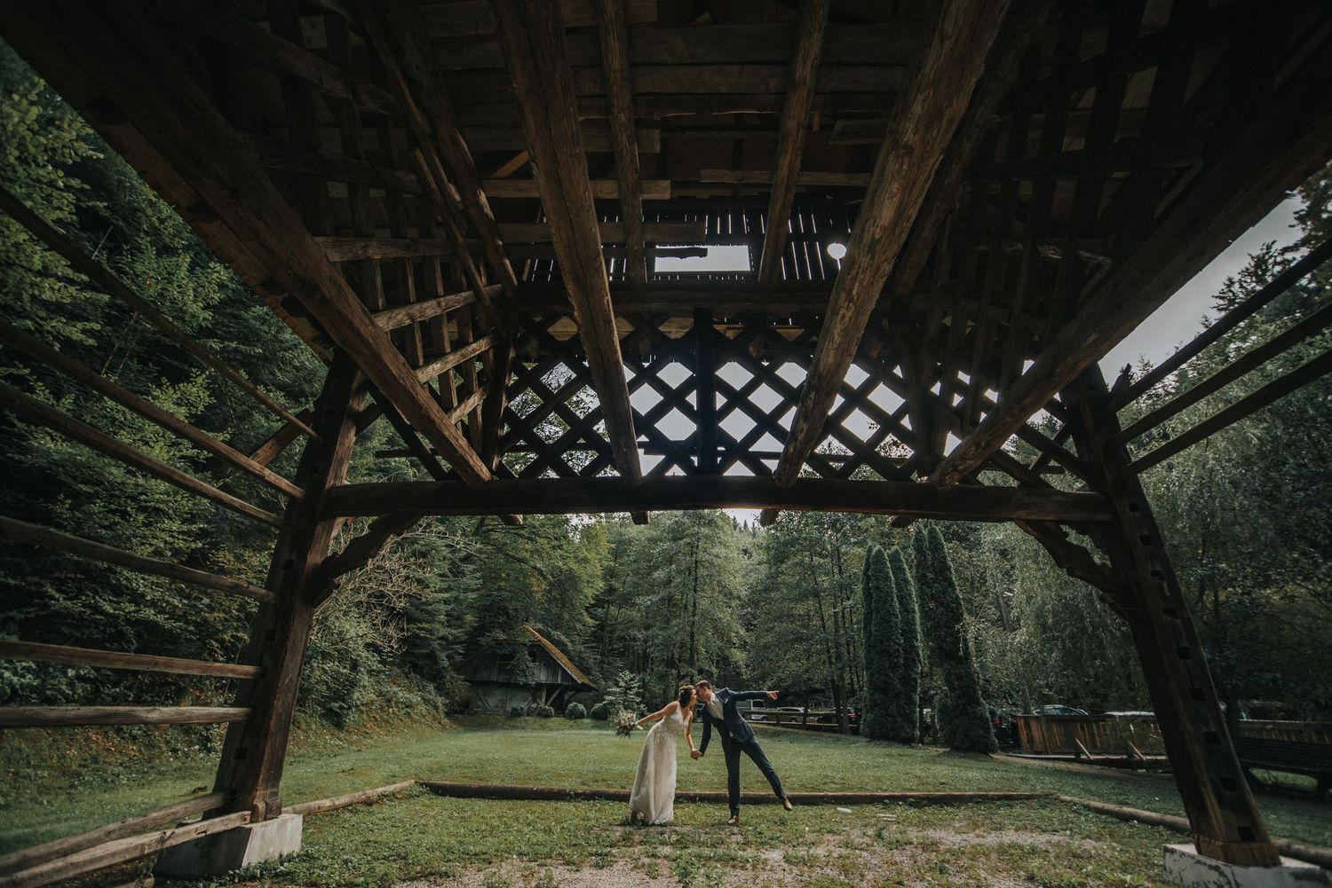 Wedding photo under wooden pavilion with forest backdrop and natural lighting.