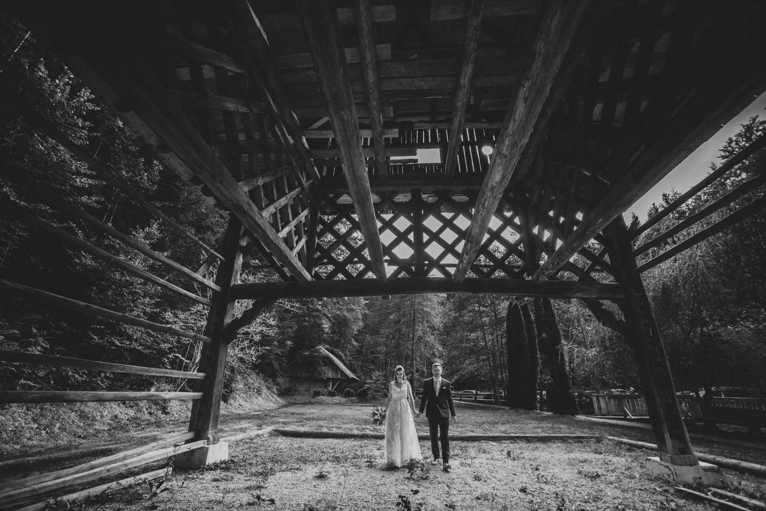 Black and white photo of couple under rustic wooden archway in forest setting.