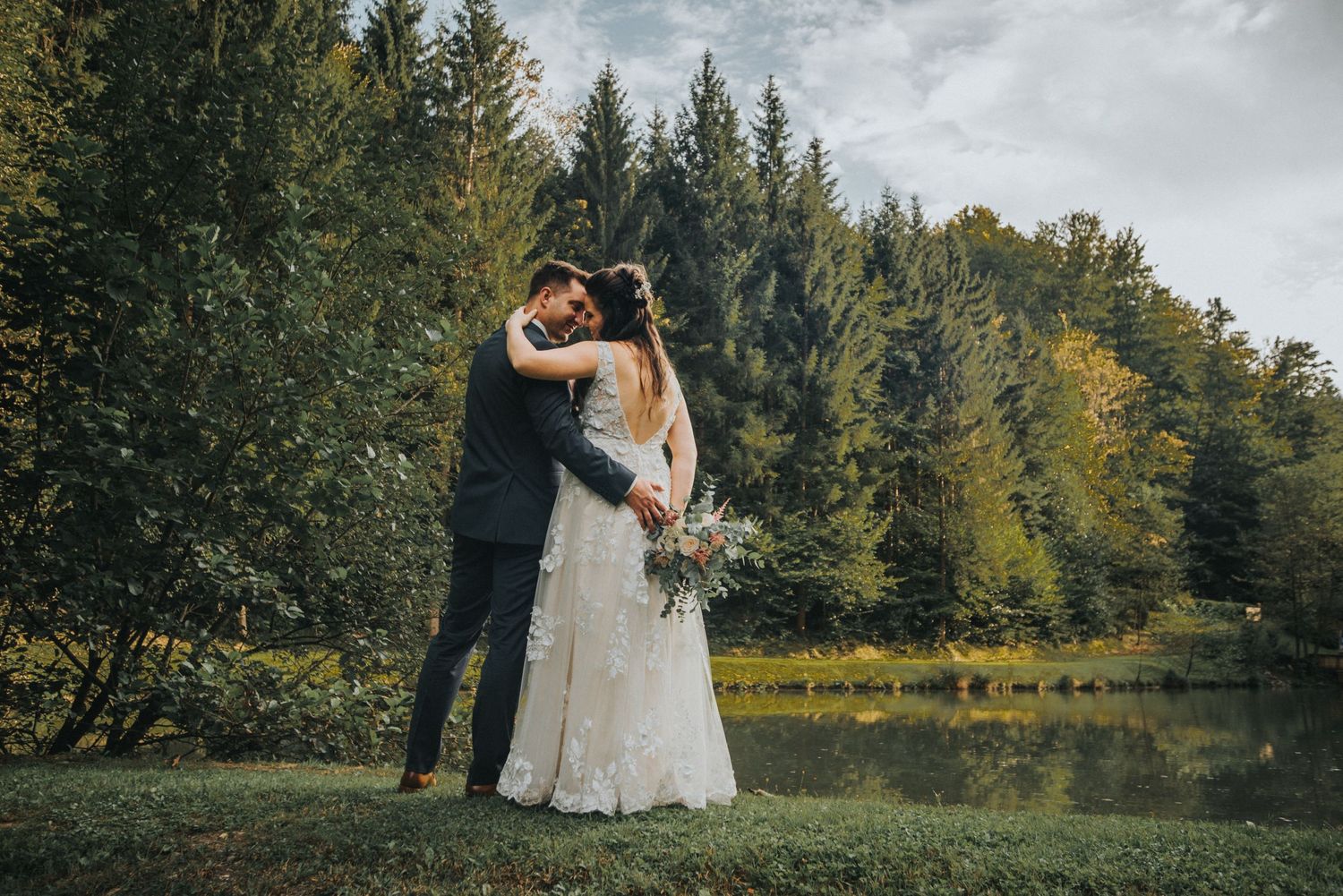A wedding couple embraces in front of a serene lake surrounded by evergreen trees and autumn foliage.