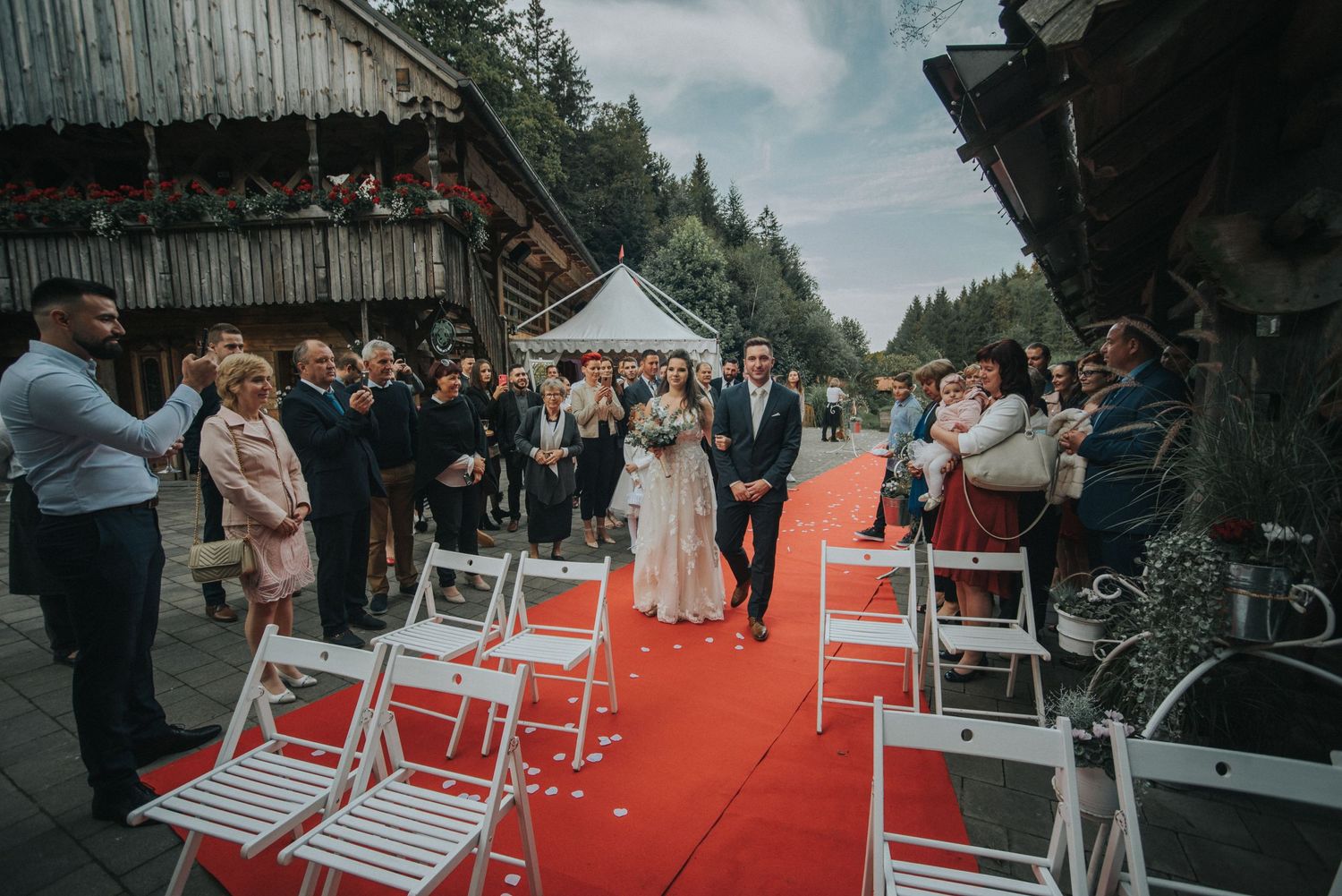 Wedding ceremony setup outdoors with red carpet aisle and white chairs surrounded by forest scenery.
