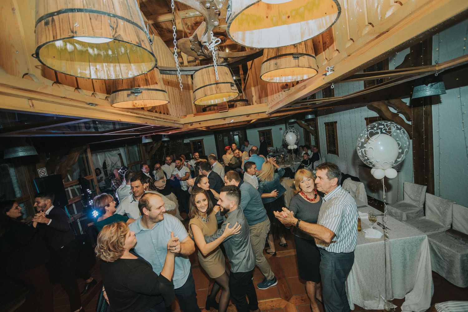 Wedding reception party scene with guests dancing under wooden ceiling with pendant lights.