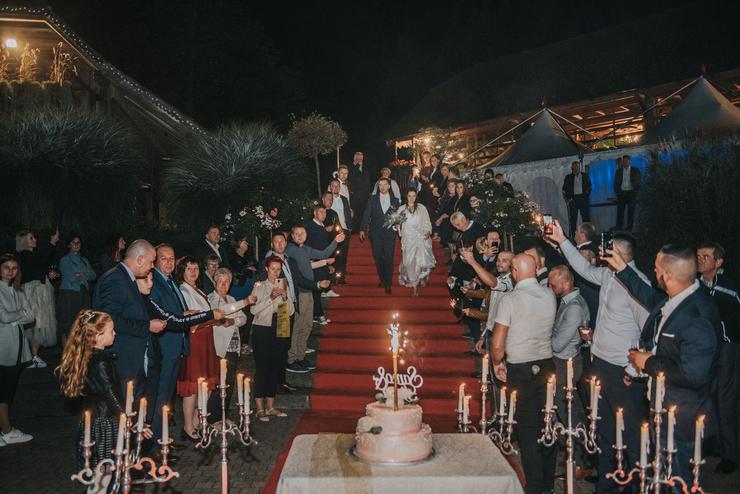 Wedding party gathers around candlelit cake table with red carpet during evening celebration.