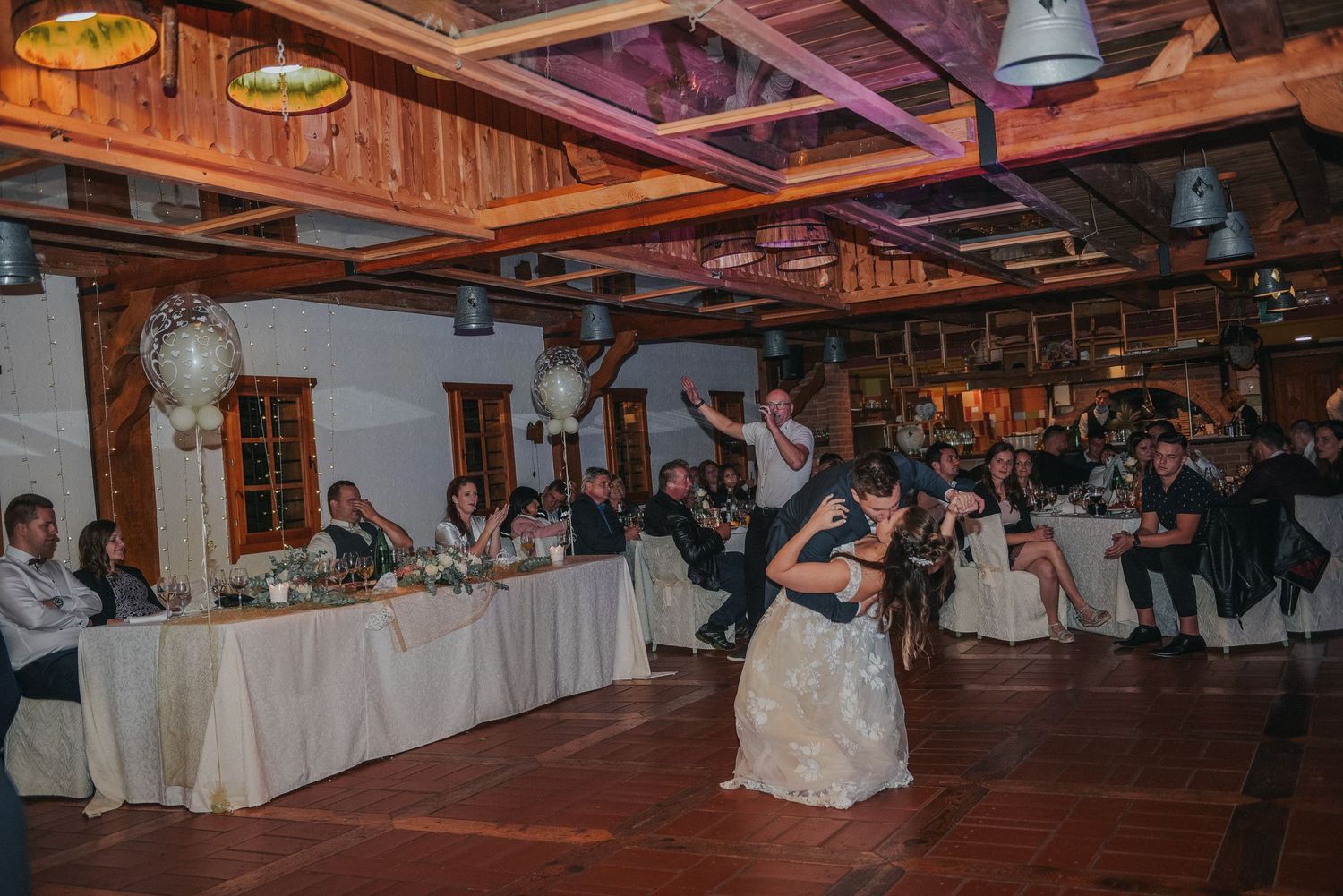Couple dancing in rustic indoor reception venue with exposed wooden beams and white tablecloths.