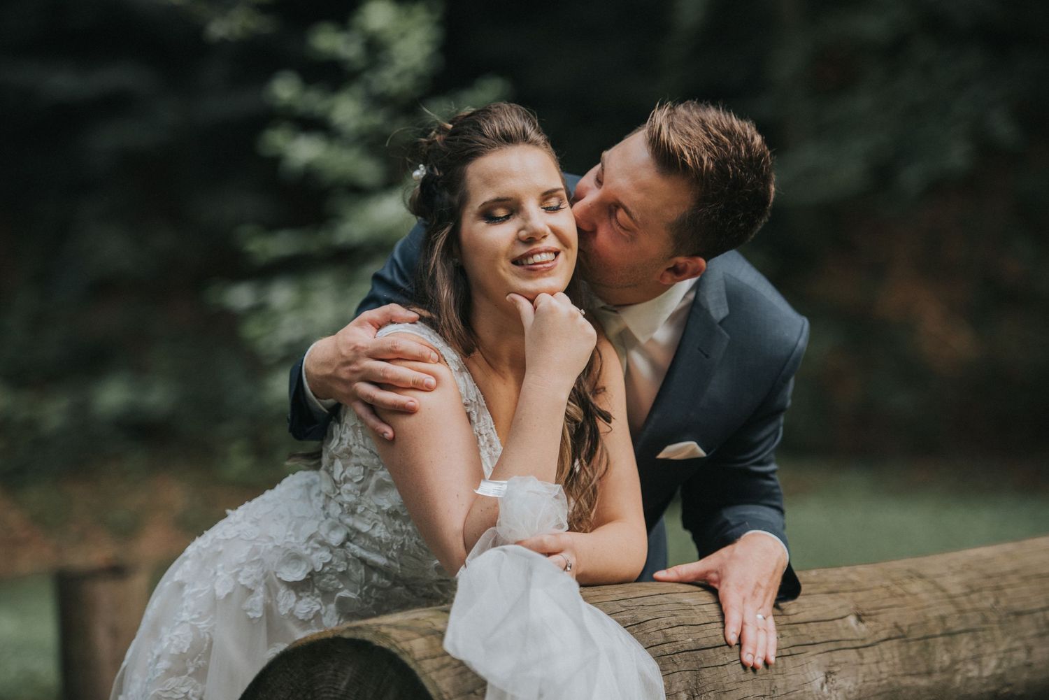 An intimate wedding moment captured on a wooden bench in a lush outdoor setting.