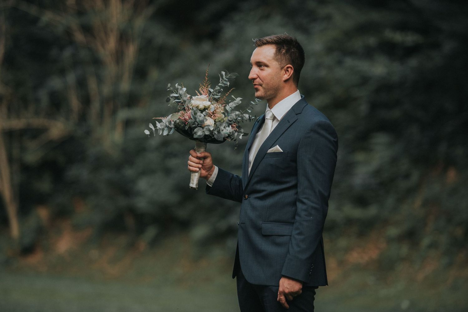 A groom in a navy suit holds a delicate bouquet of flowers against a blurred forest backdrop.