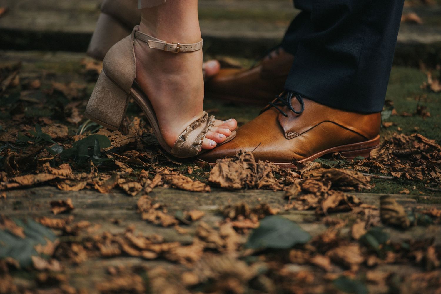 Close-up of bride and groom's feet among fallen autumn leaves, showing elegant wedding shoes.