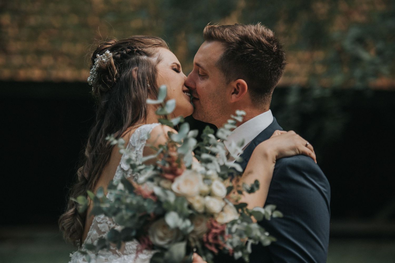 A romantic moment with a wedding bouquet featuring soft neutral flowers against a brick wall. Hiša Lisjak