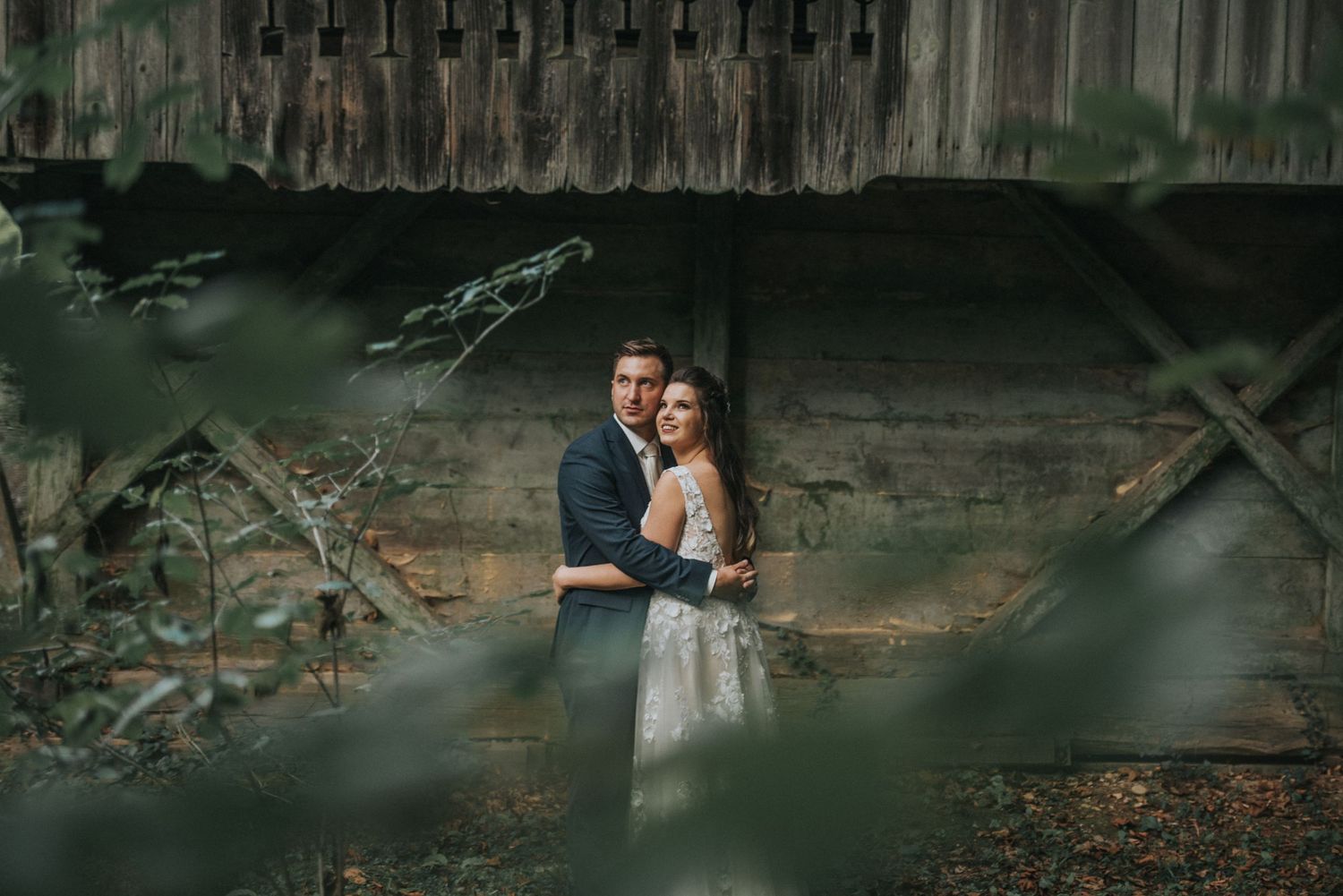 A dreamy wedding portrait through foliage against a rustic wooden backdrop.