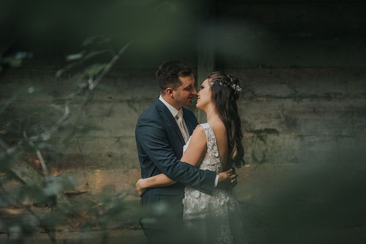 A romantic wedding couple embraces in a moody outdoor setting with soft bokeh and natural elements.