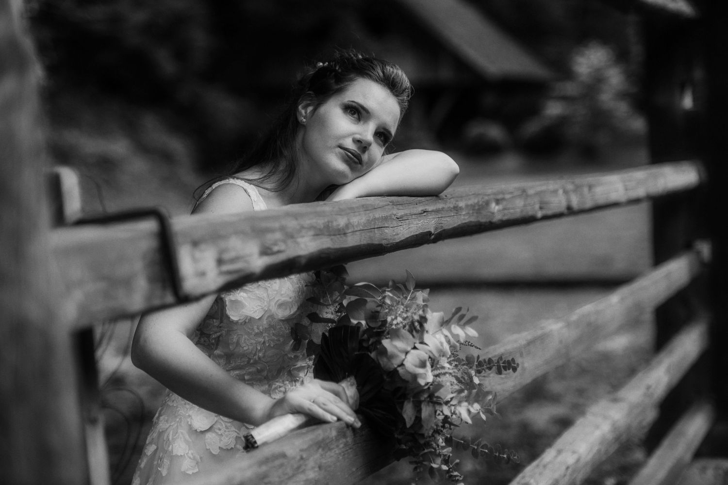 A dreamy black and white portrait of a bride holding flowers while leaning on a wooden fence.