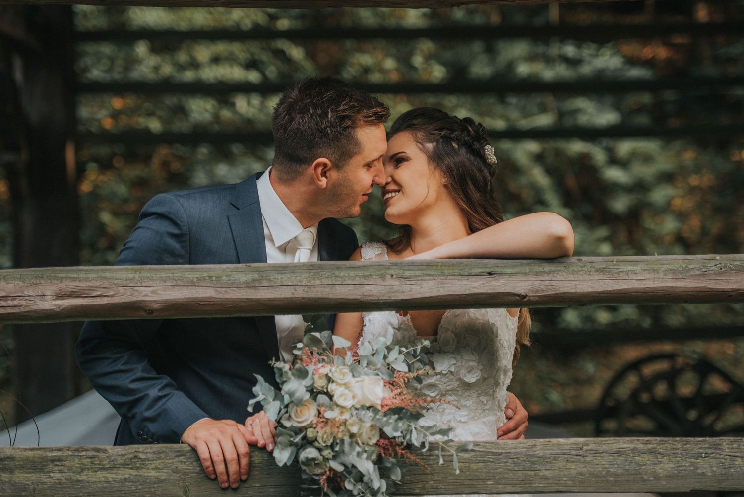 Newlyweds share an intimate moment on a rustic wooden bench with a beautiful floral bouquet.