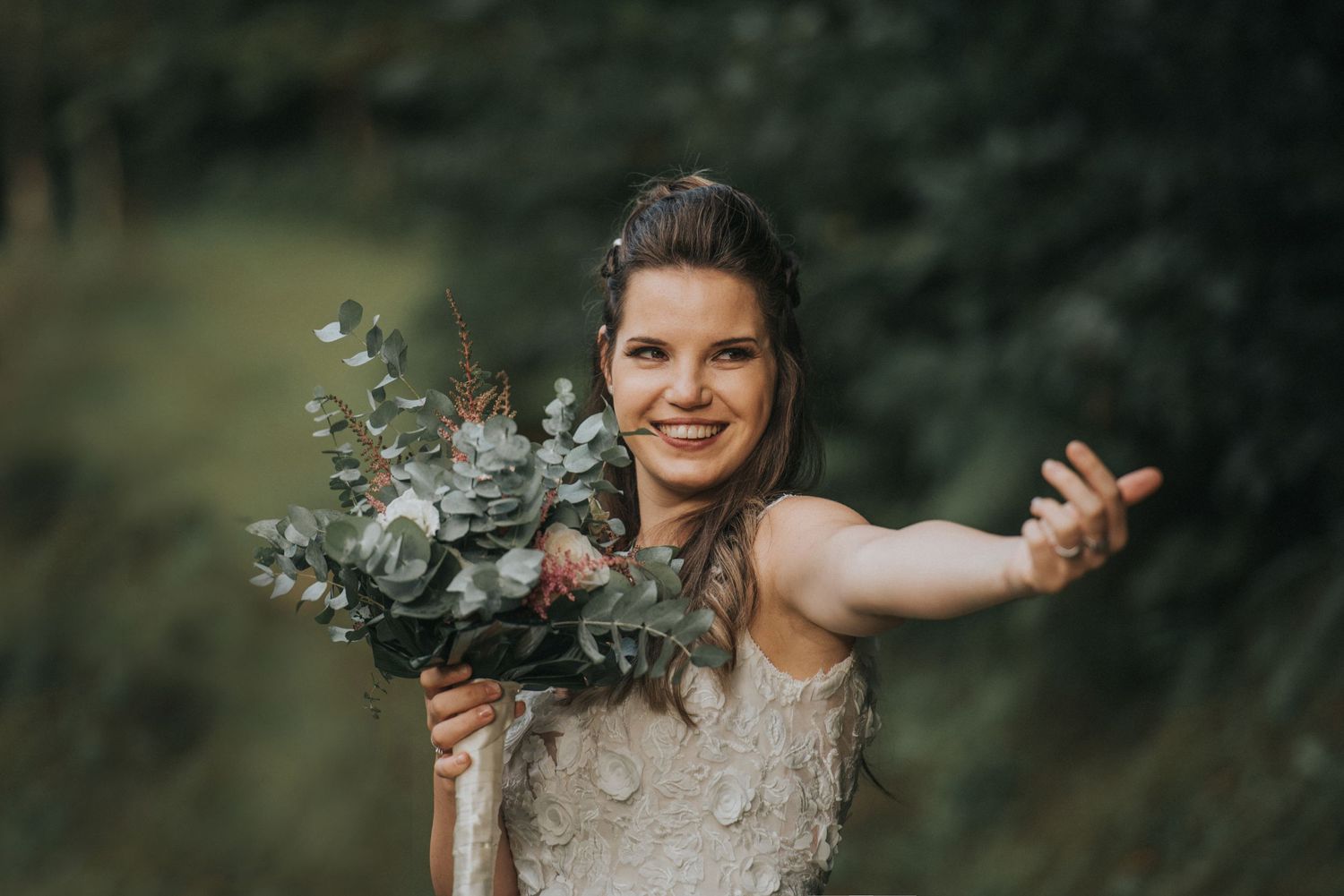 A joyful bride holding a floral bouquet with arms spread wide in a natural outdoor setting.