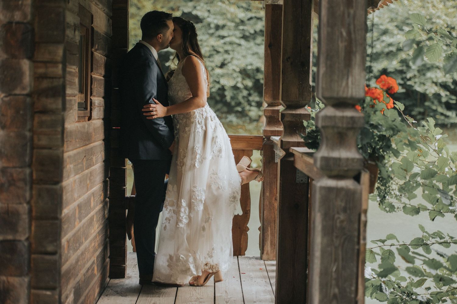 A romantic moment between newlyweds sharing a kiss on a rustic wooden porch with flowers in the background.