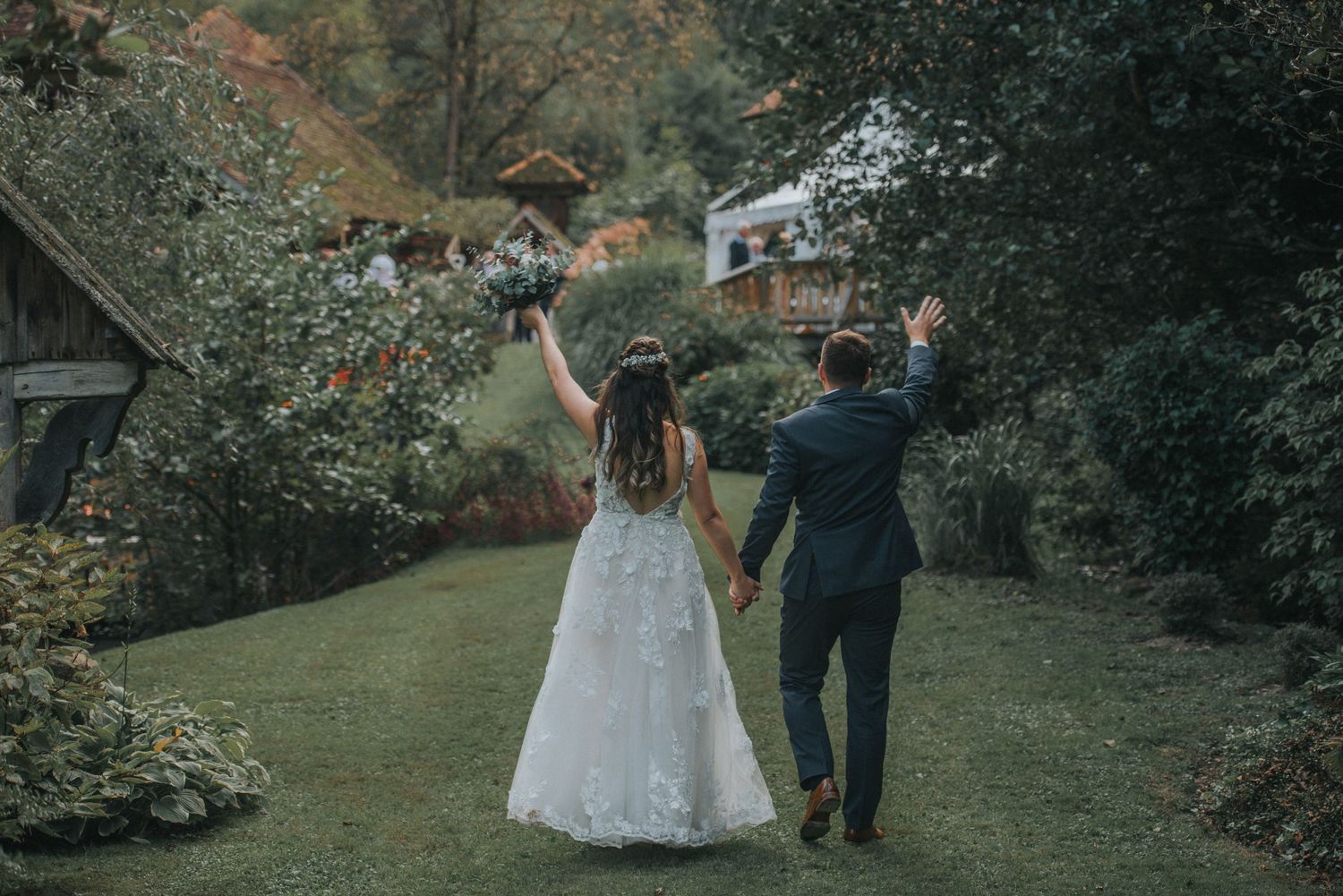 A wedding couple celebrates on a garden path while walking away from the camera with raised hands.