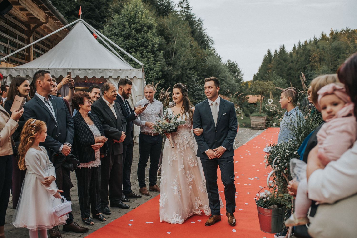 Outdoor wedding ceremony on red carpet with forest backdrop and white tent.