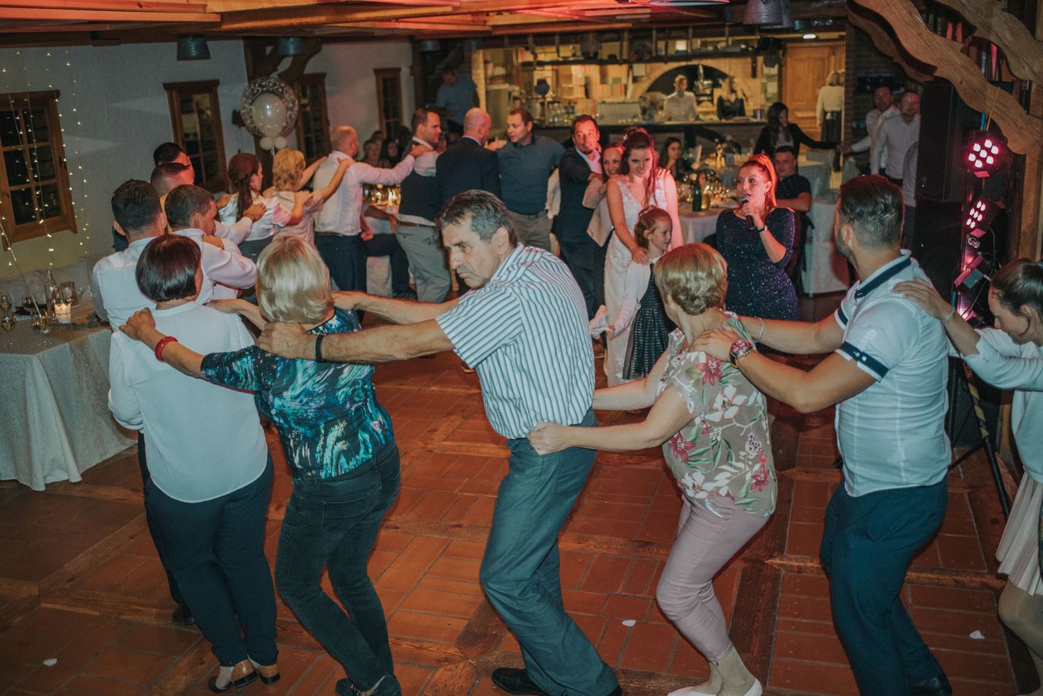 Large group of wedding guests dancing in a circle at indoor reception venue.