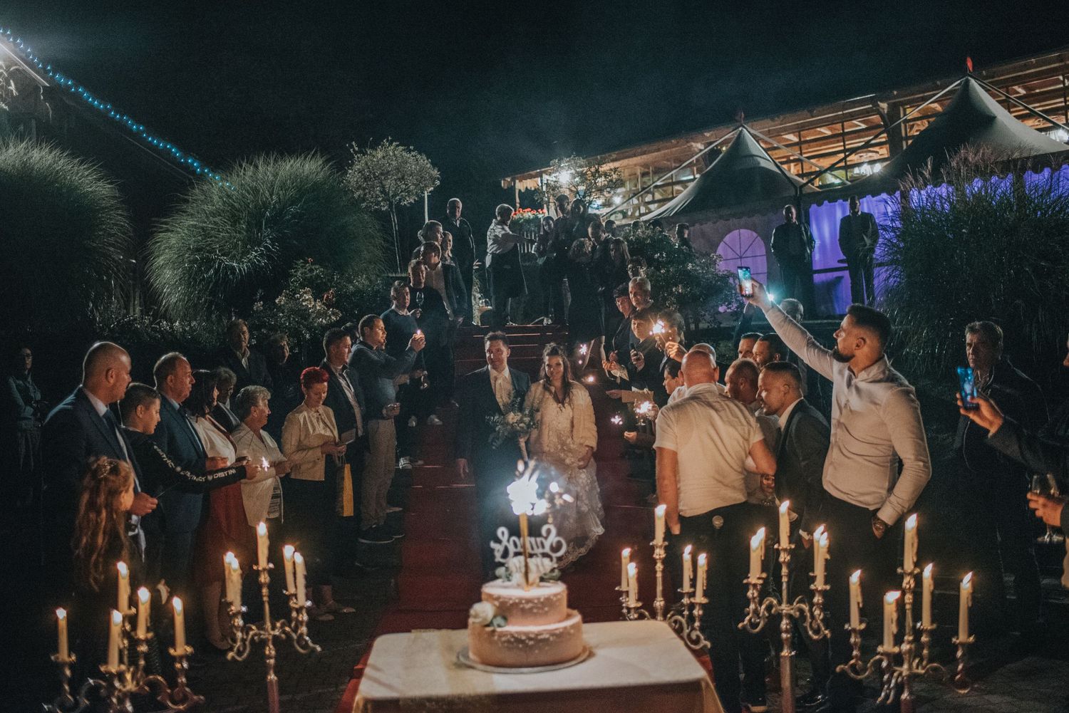 Wedding celebration at night with guests gathered around illuminated cake and candles.