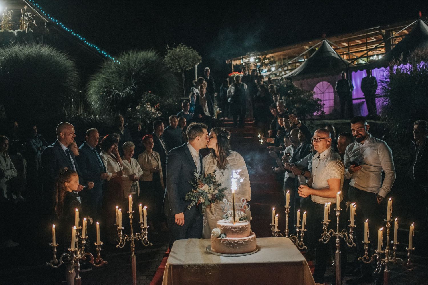 Nighttime wedding cake cutting ceremony surrounded by guests holding lit candles.