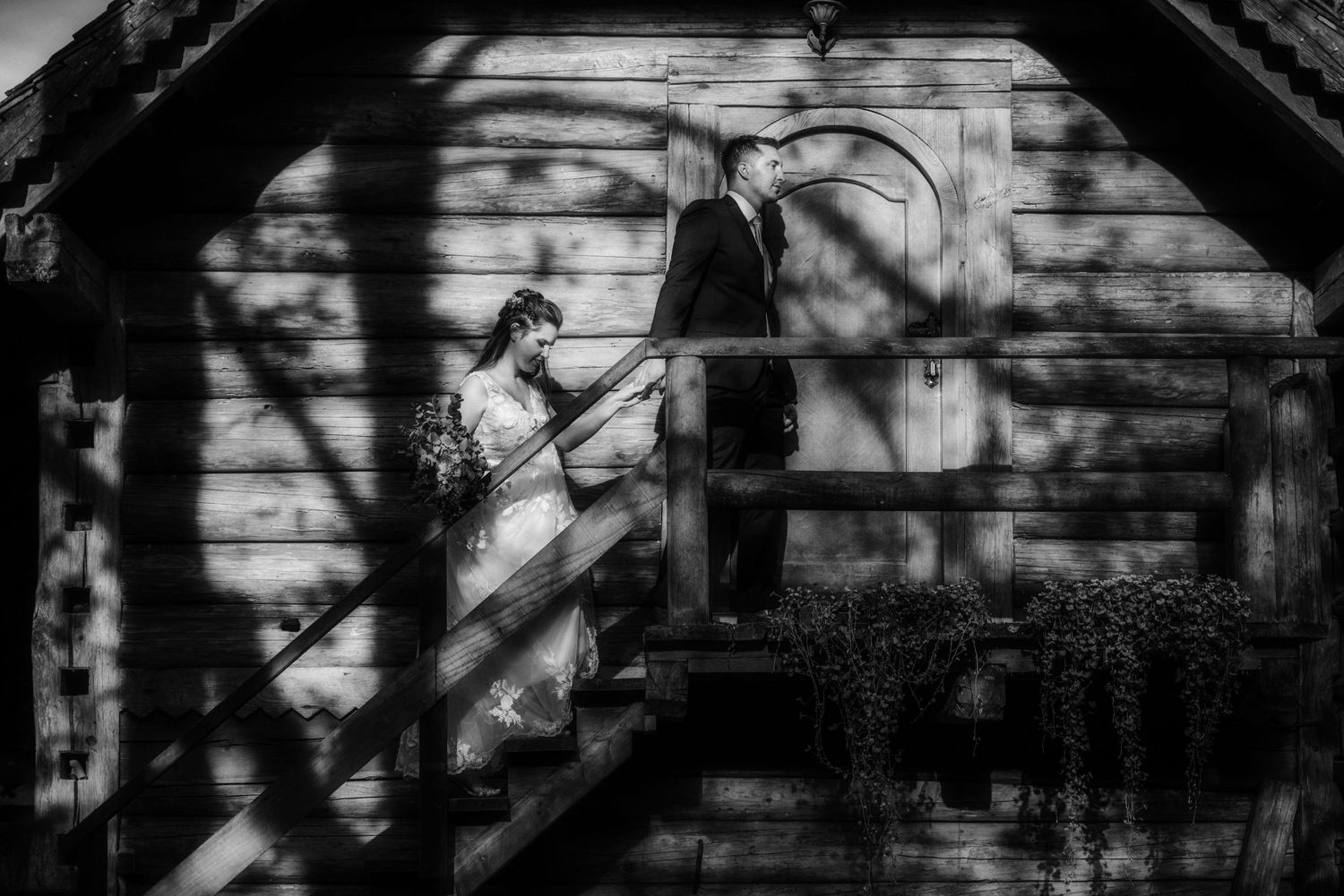 Black and white photo of a figure ascending stairs in dappled sunlight against a rustic wooden building.