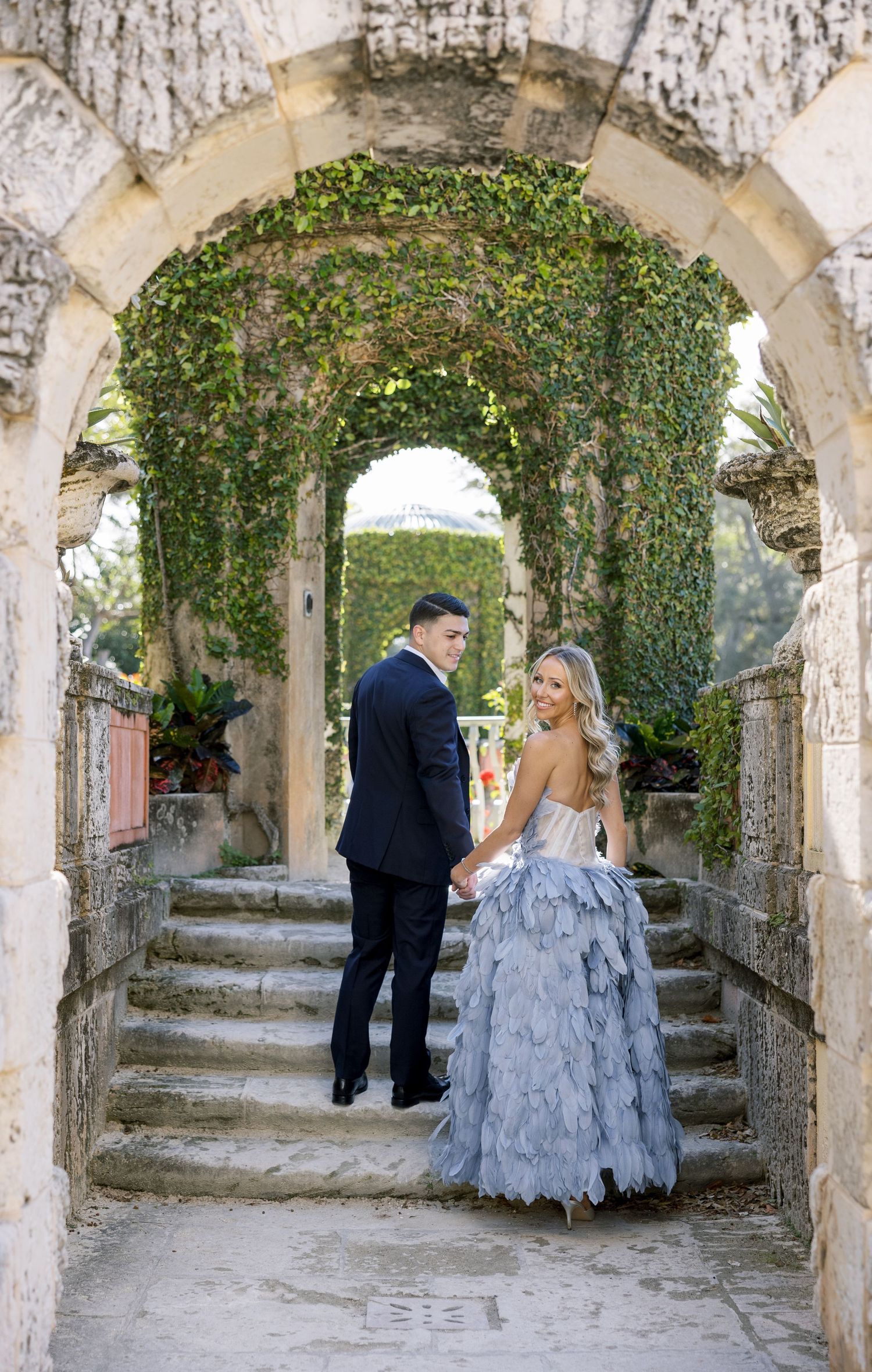 A couple stands in a vine-covered archway, with the woman wearing a blue ruffled gown.