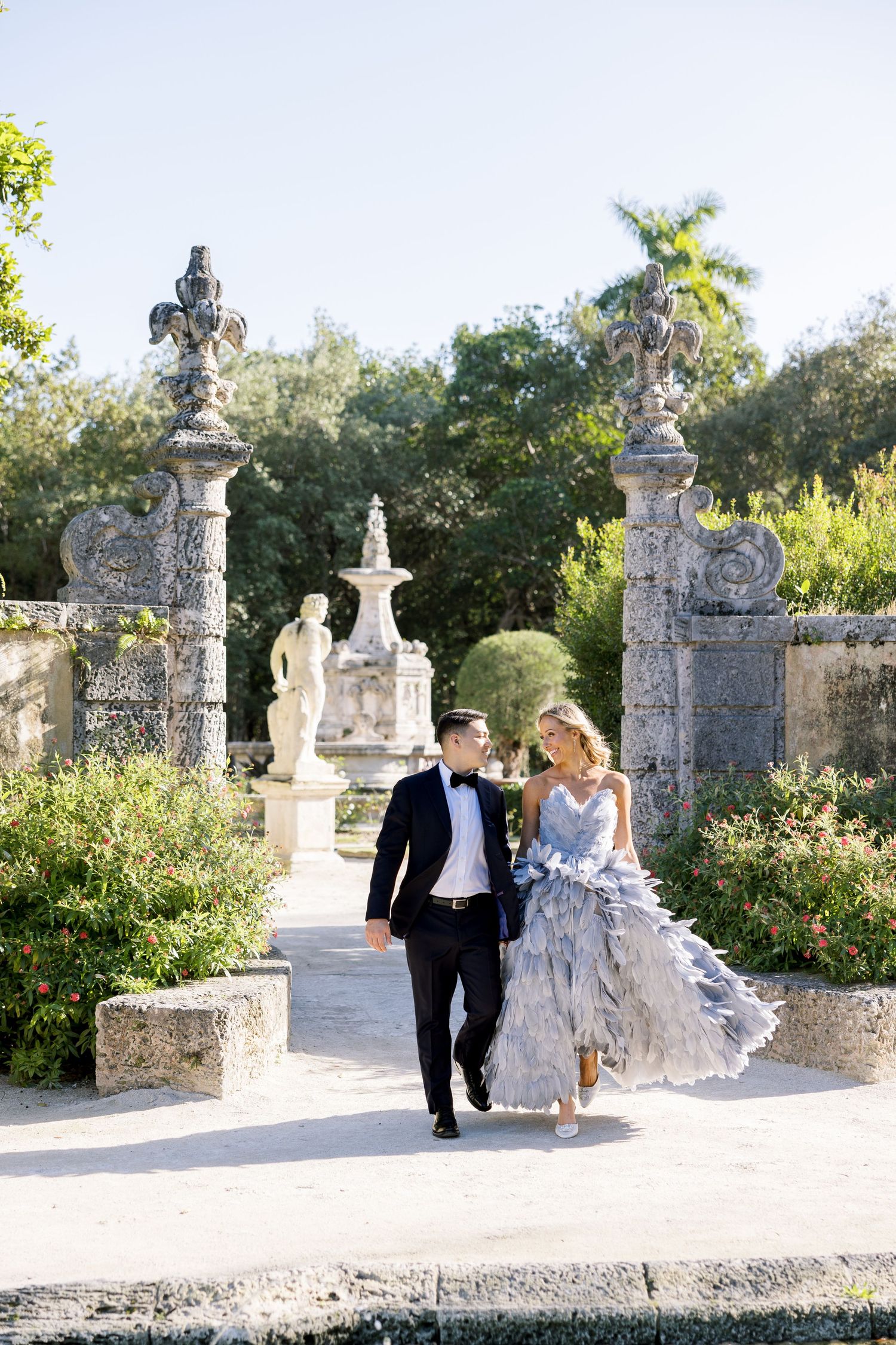 A couple strolling through an ornate garden walkway with classical stone pillars and sculptures.
