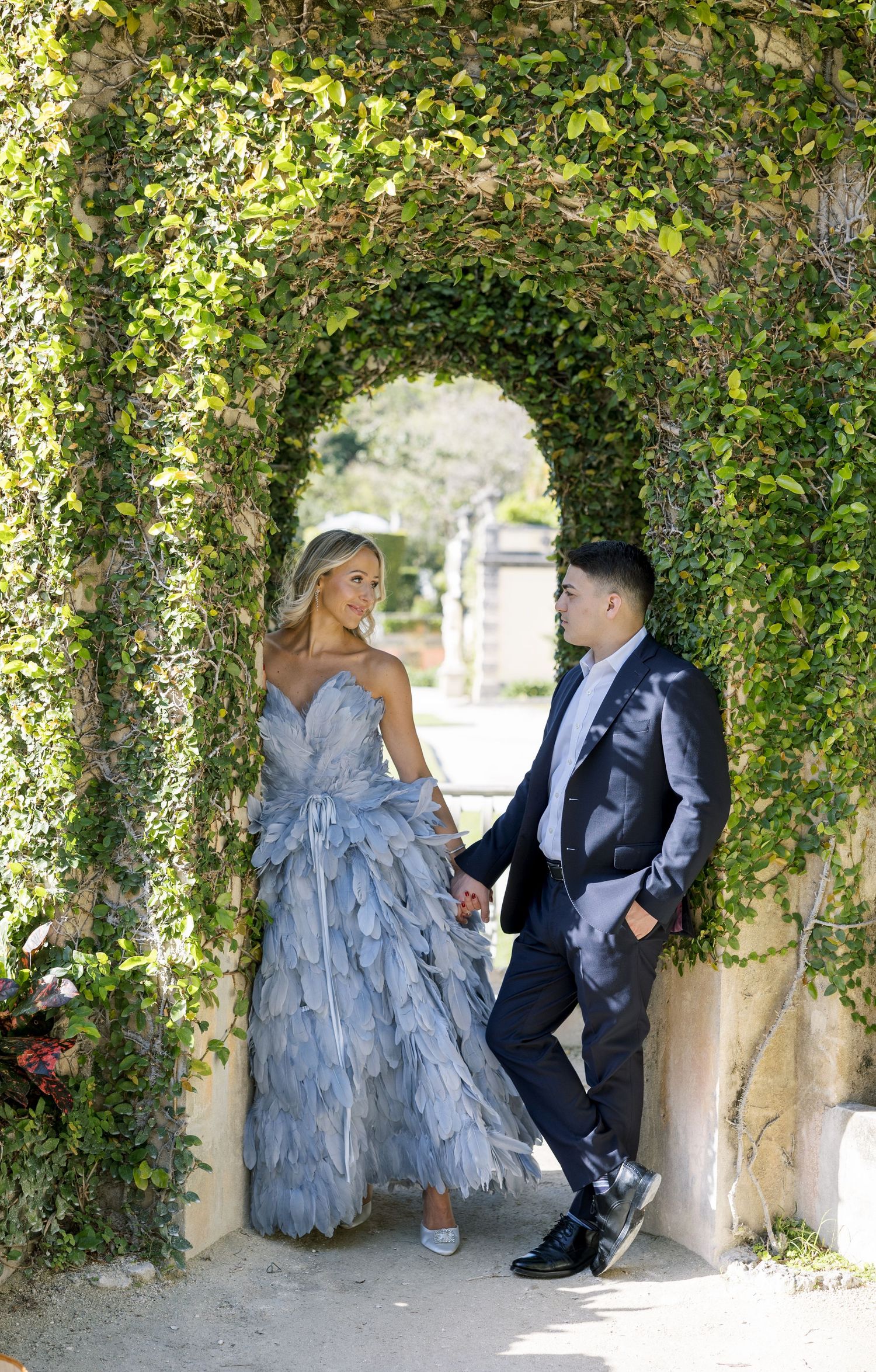 A couple poses in formal attire beneath a lush green garden archway, with the bride wearing a blue feathered gown.