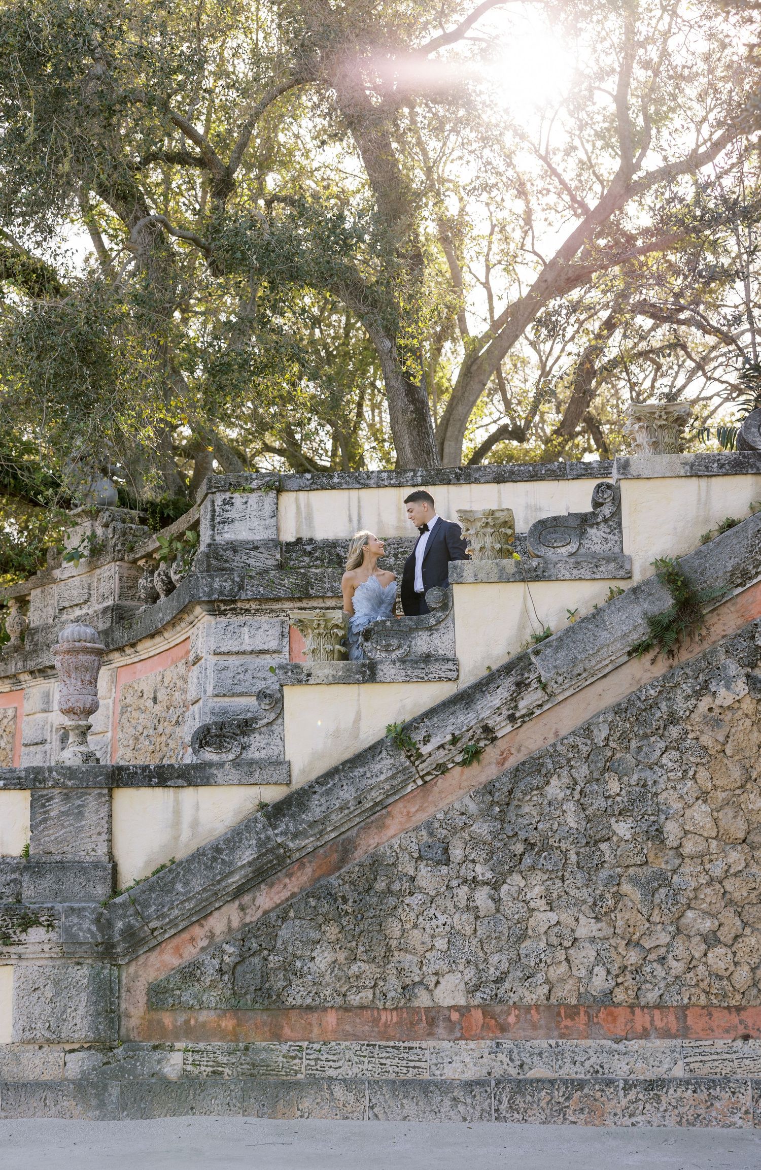 A romantic photoshoot on grand stone steps surrounded by lush trees and sunlight.