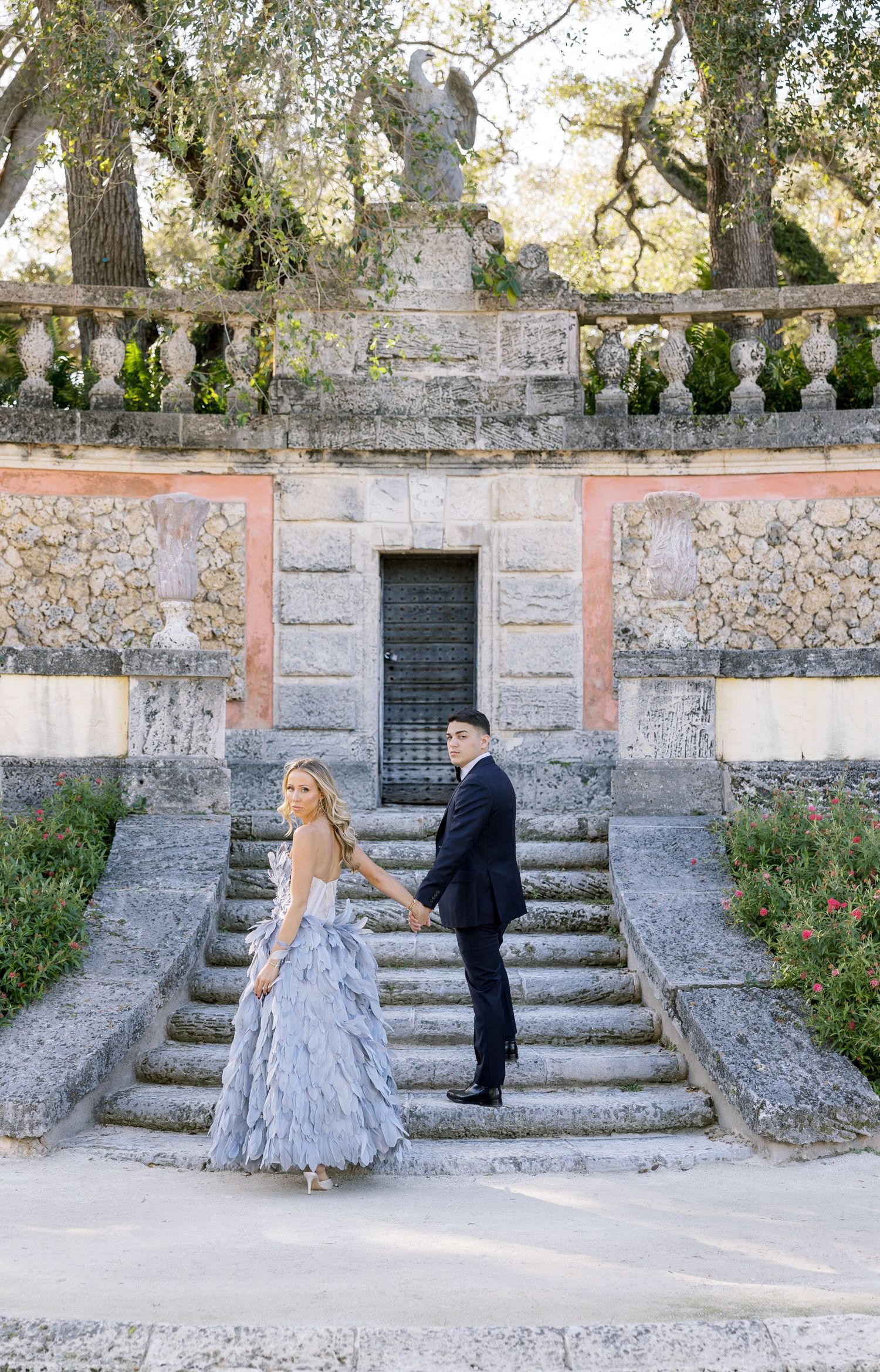 Couple in formal attire standing on historic stone steps with ornate balustrades and garden architecture.