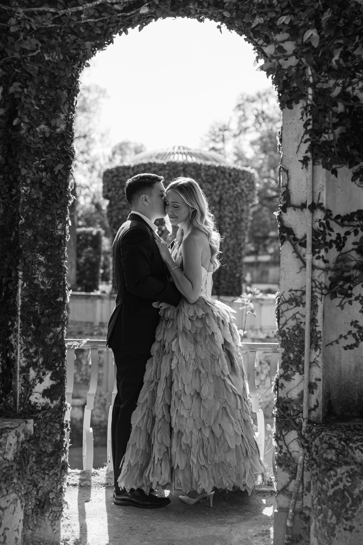 A romantic black and white wedding photo of a couple embracing under an ivy-covered archway in a garden setting.