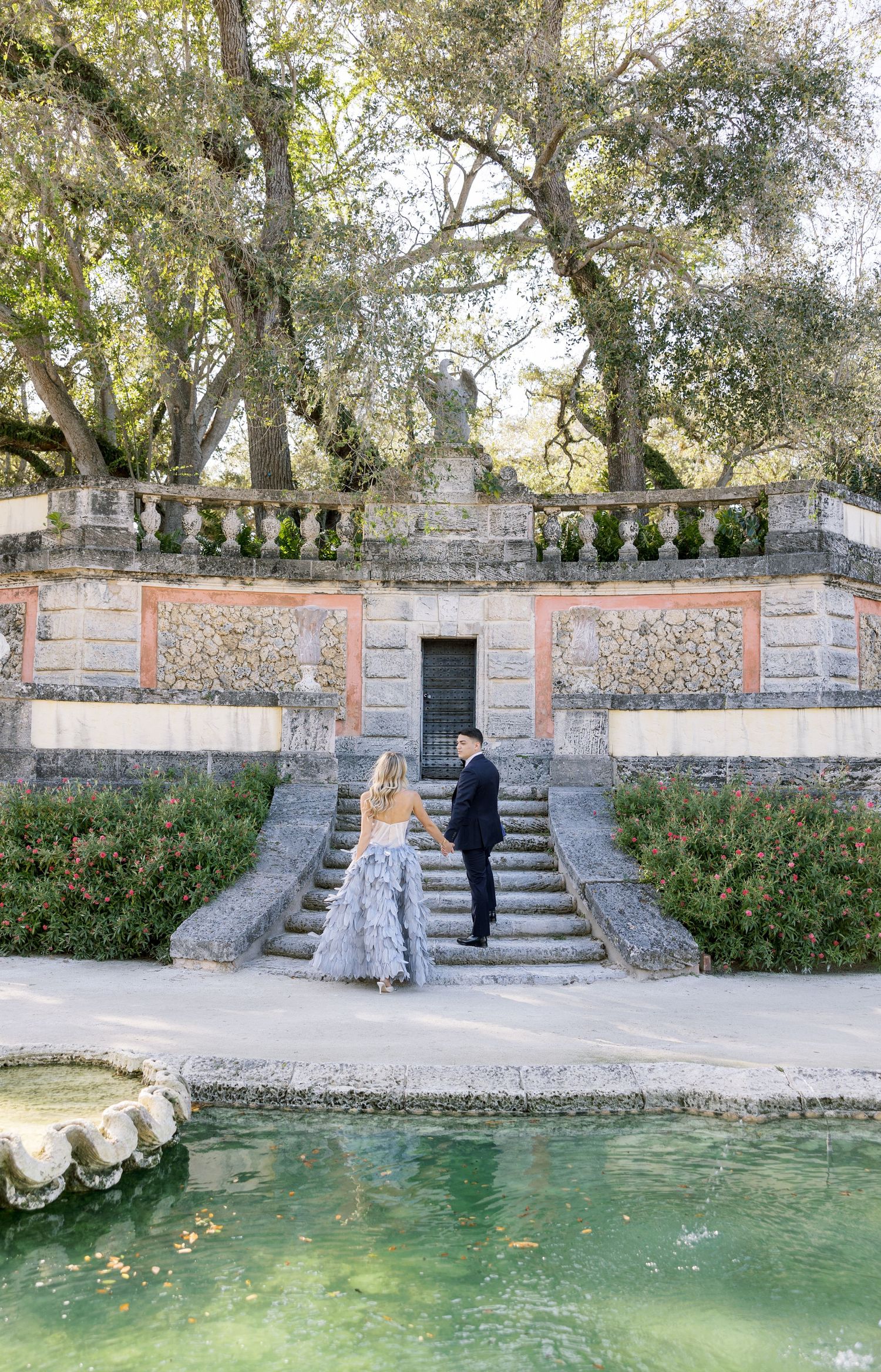 Couple in formal wear standing on stone steps overlooking a reflecting pool in a formal garden.