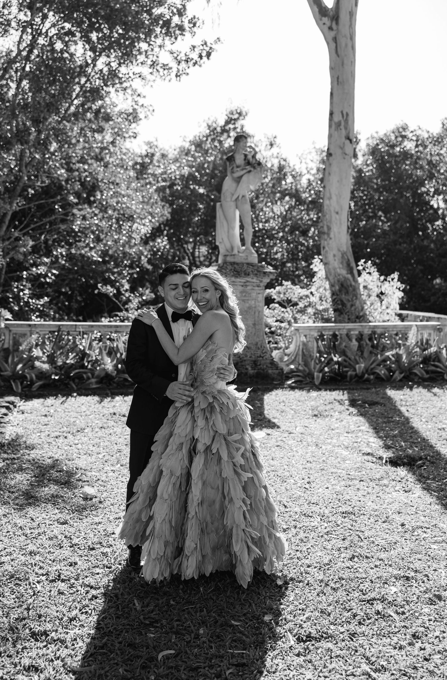 Black and white portrait of a couple embracing in a formal garden with classical statue in background.