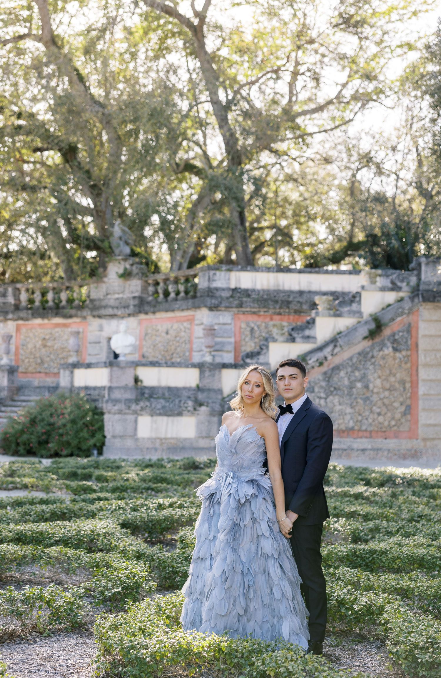 Formal portrait of a couple on garden terraces with brick walls and trees in background.