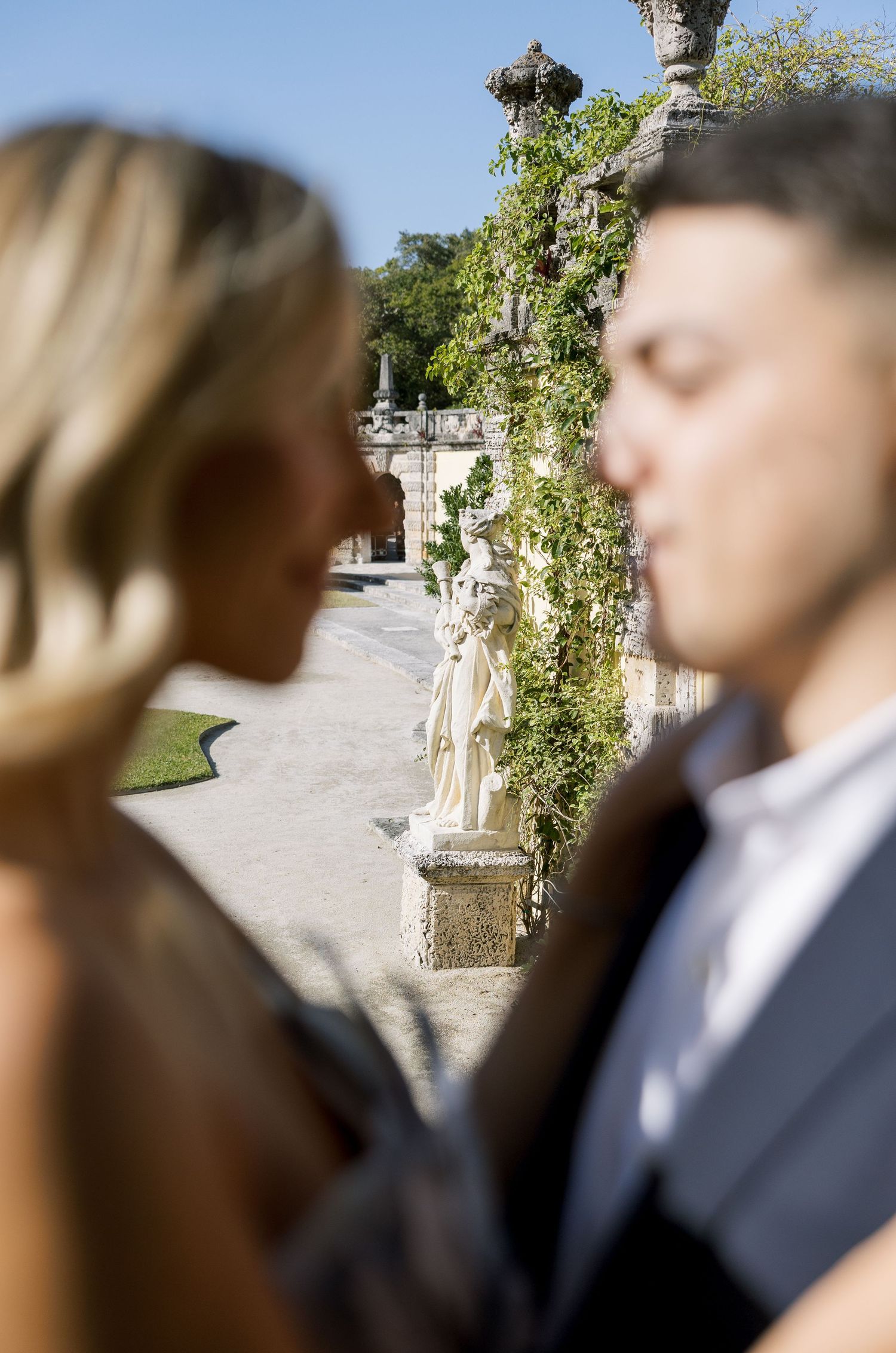 Close-up portrait of a couple with garden statues and architecture in soft focus background.