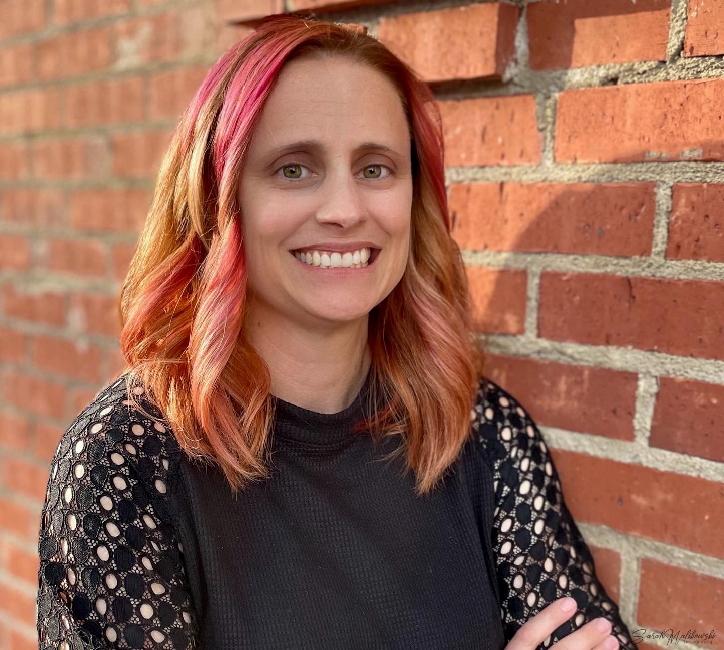 Portrait against red brick wall showing vibrant pink-tinted hair and black lace sleeves.