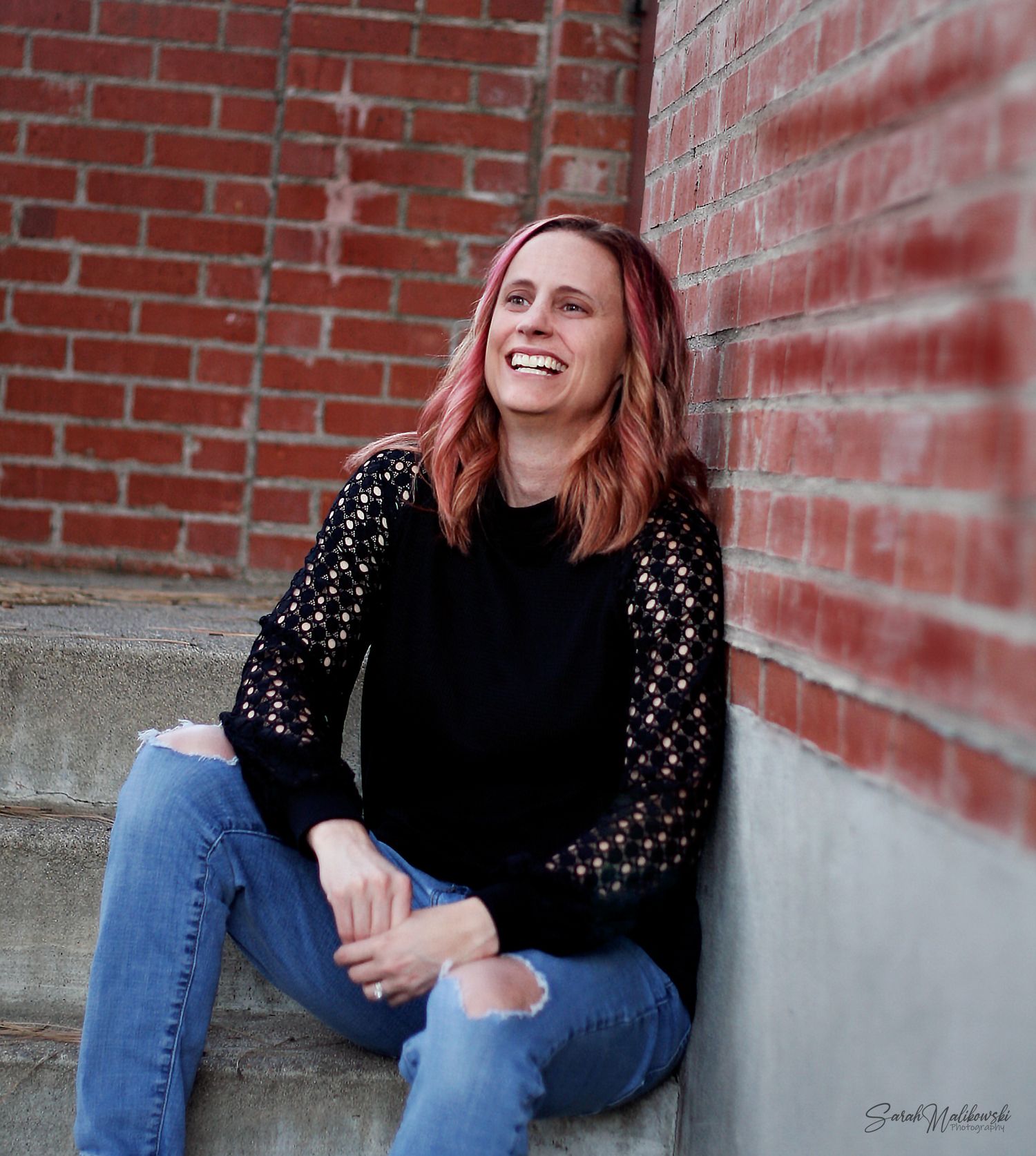 Candid portrait by brick wall wearing black sweater with patterned sleeves and distressed jeans.
