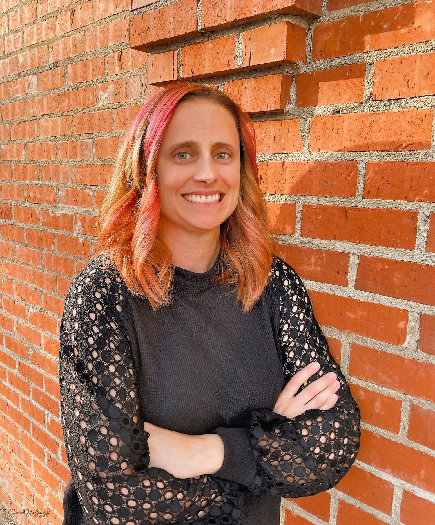 Portrait series against orange brick featuring someone in a black top with lace sleeves.